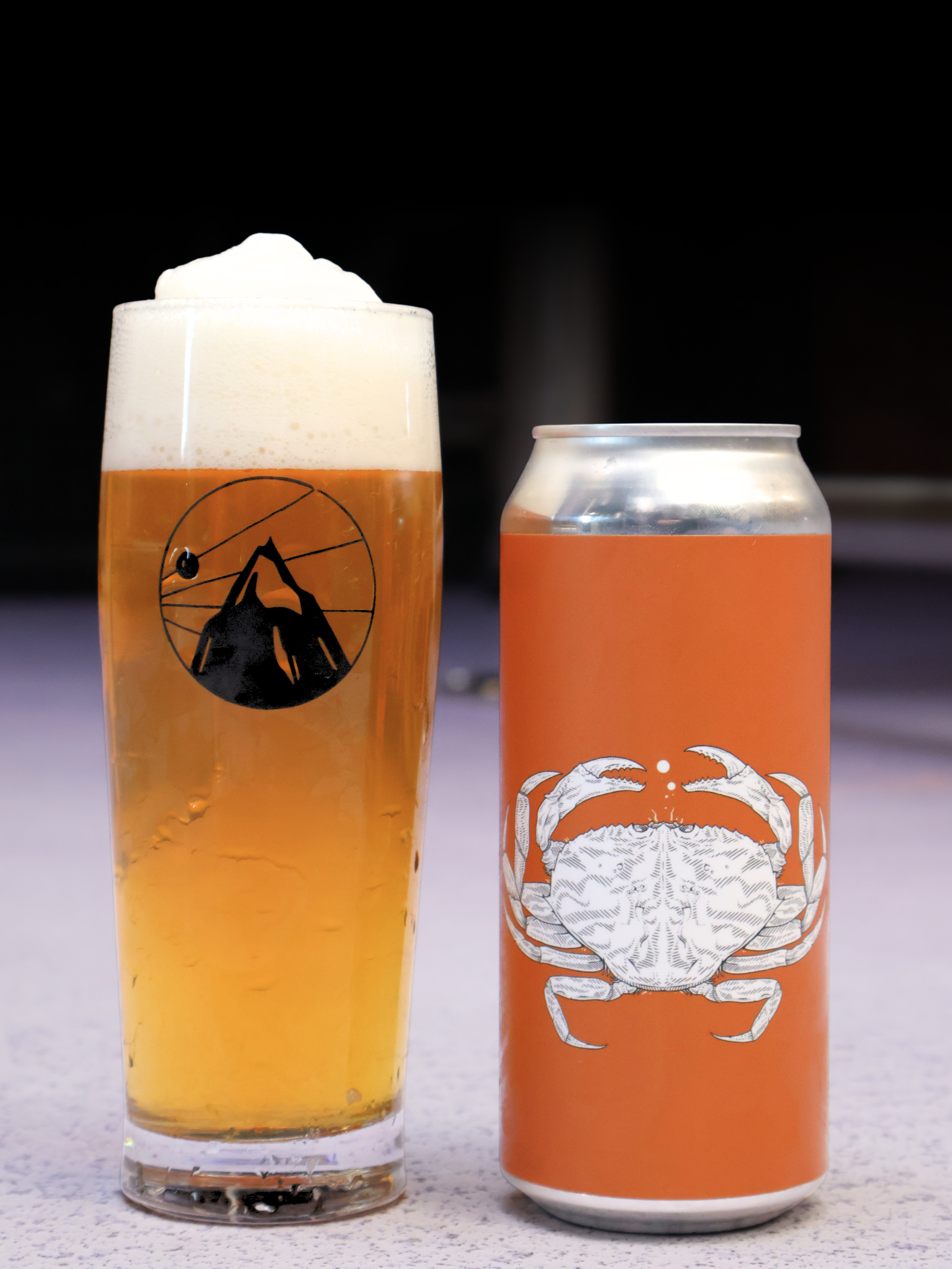 A glass of beer with a mountain and sun logo, topped with foam, and an orange soda can with a crab illustration, on a light-colored surface.