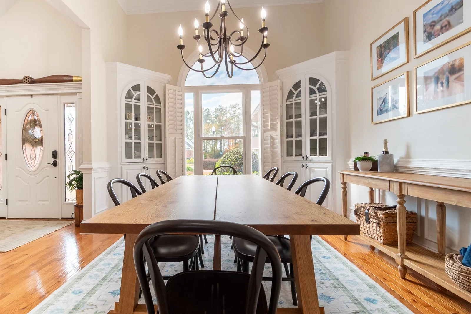 A dining room with a wooden table surrounded by black chairs, a chandelier hanging from the ceiling, large windows with shutters, a sideboard with decorations, and framed photos on the wall.