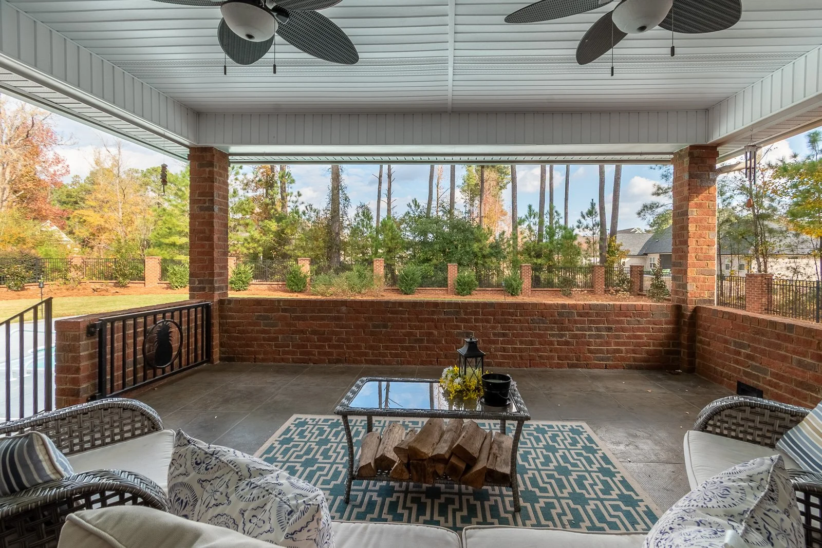 Covered outdoor patio with brick walls, ceiling fans, and a sitting area including a sofa, chairs, a glass-top table, and a decorative rug, overlooking a yard with trees and a fence in the background.