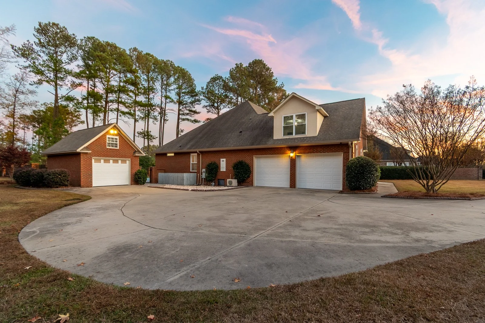 A large brick house with two white garage doors, a smaller detached brick garage, and a driveway. The house has a second-story window and is surrounded by trees, with a sunset sky in the background.