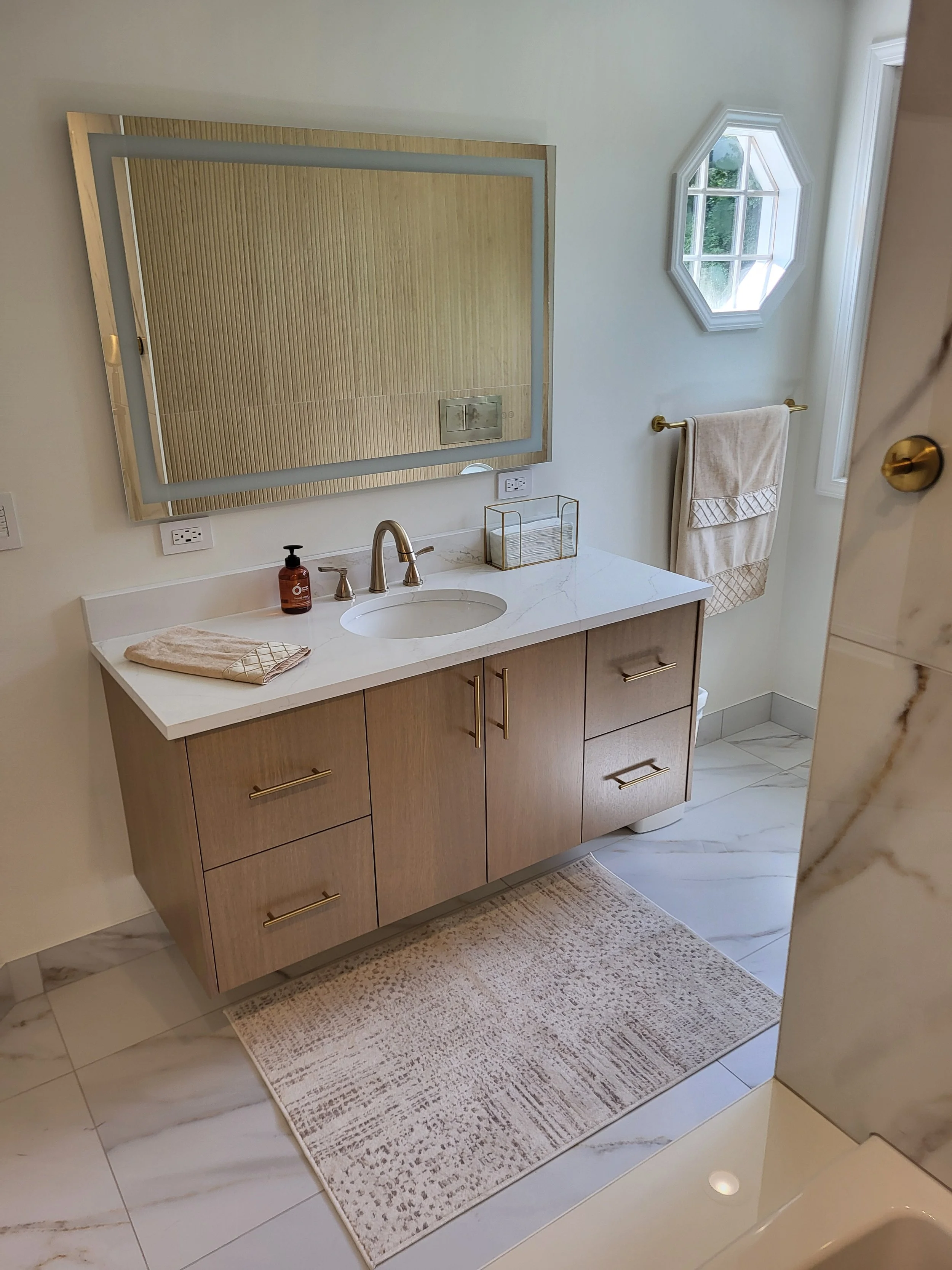 Bathroom with a wooden vanity, marble countertop, large mirror, octagonal window, beige towels, and a patterned rug on marble floor.