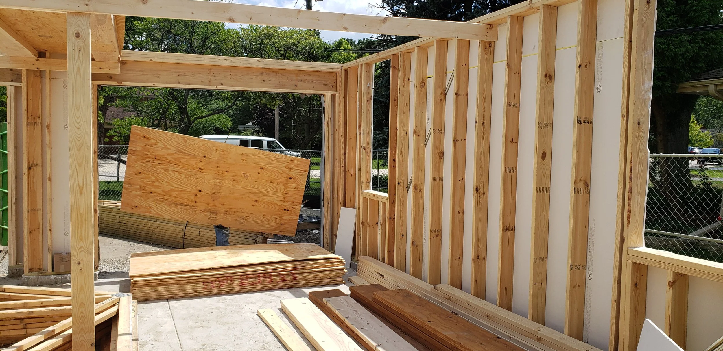 Construction site with wooden framing and plywood sheets, partially built wall, outdoor setting with trees and a fence in the background.