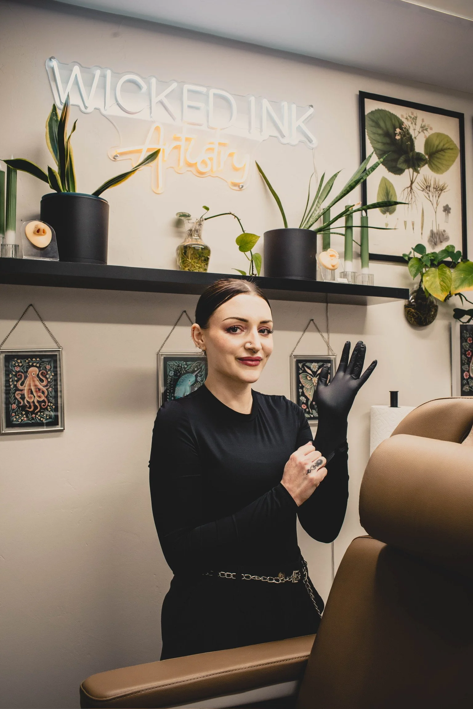 A woman with dark hair, wearing black gloves and a black top, is standing in a tattoo studio. She is smiling, adjusting her gloves, and standing next to a chair. Behind her, there's a wall with framed artwork and a sign that reads "Wicked Ink Tattoo" in neon lights. The studio interior includes potted plants and decorative art.