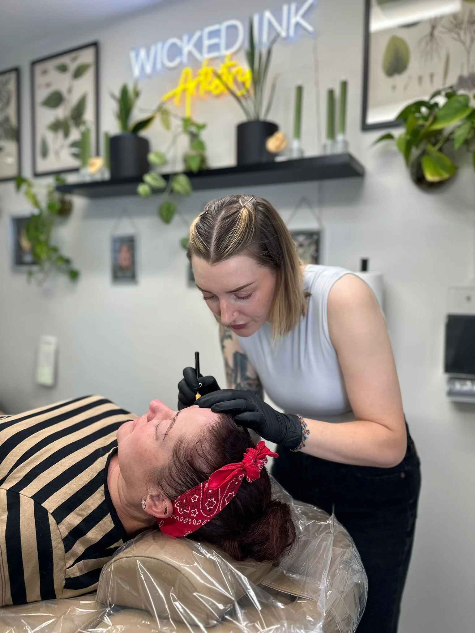A woman is laying on a treatment table receiving permanent makeup tattooing on her eyebrows from a professional artist in a modern studio with plant decor.