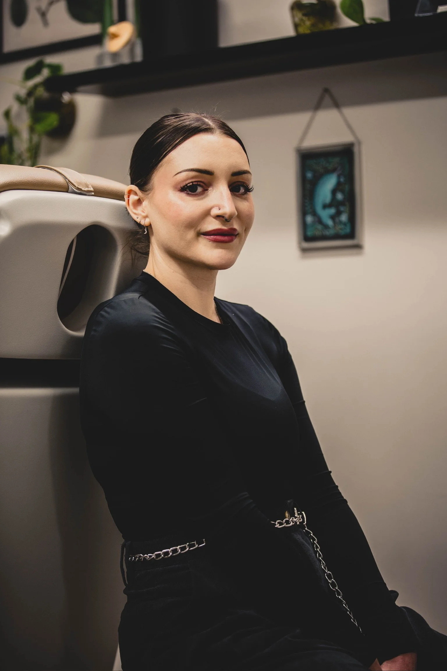 A woman with dark hair and makeup, sitting against a white salon sink, wearing a black top and silver chain belt, with decorative wall art and plants in the background.