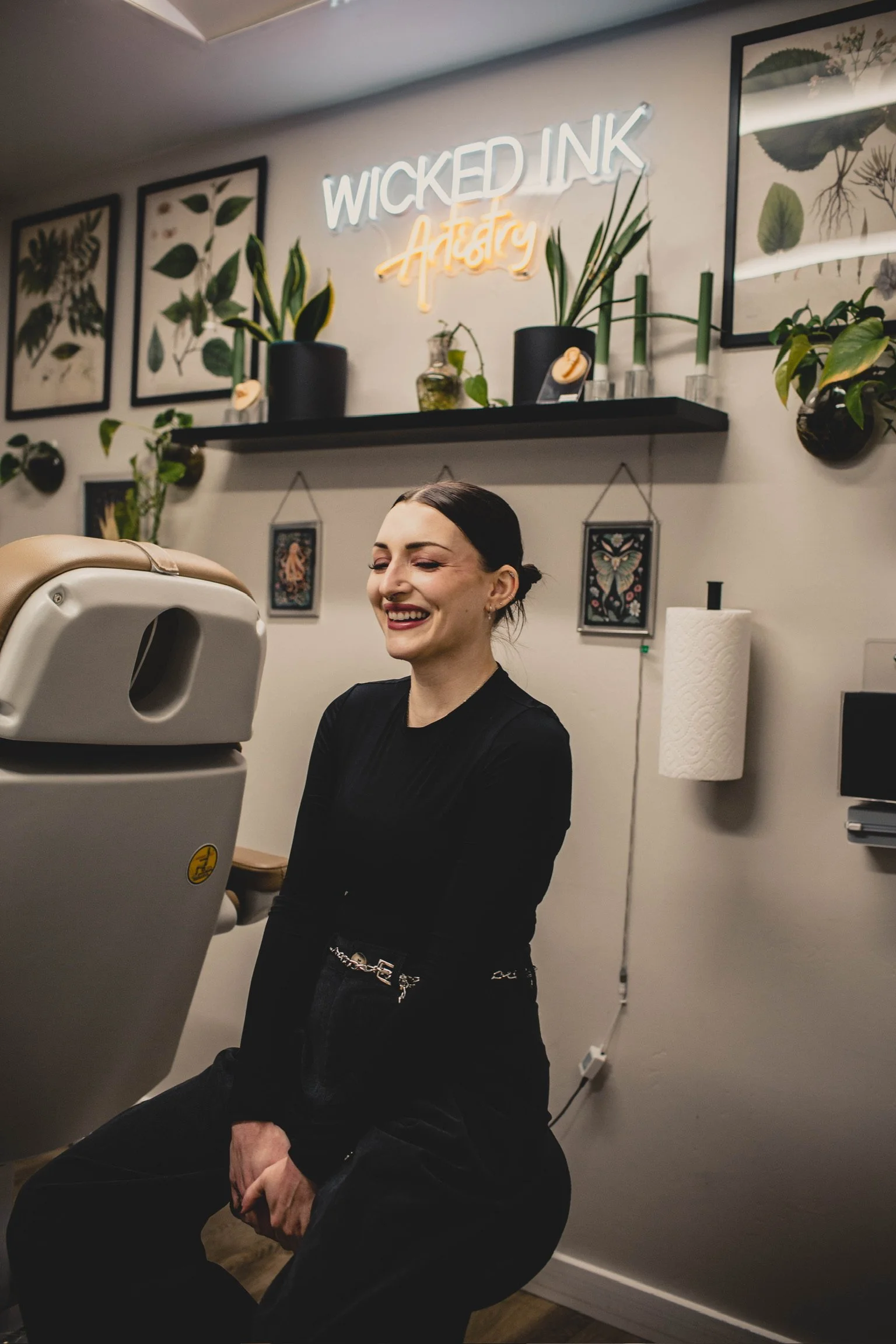 A smiling woman in black clothing sitting in a tattoo studio, with tattoo equipment partially visible in front of her. The studio features framed botanical artwork, potted plants, and a neon sign that reads 'WICKED INK Artistry' on the wall.
