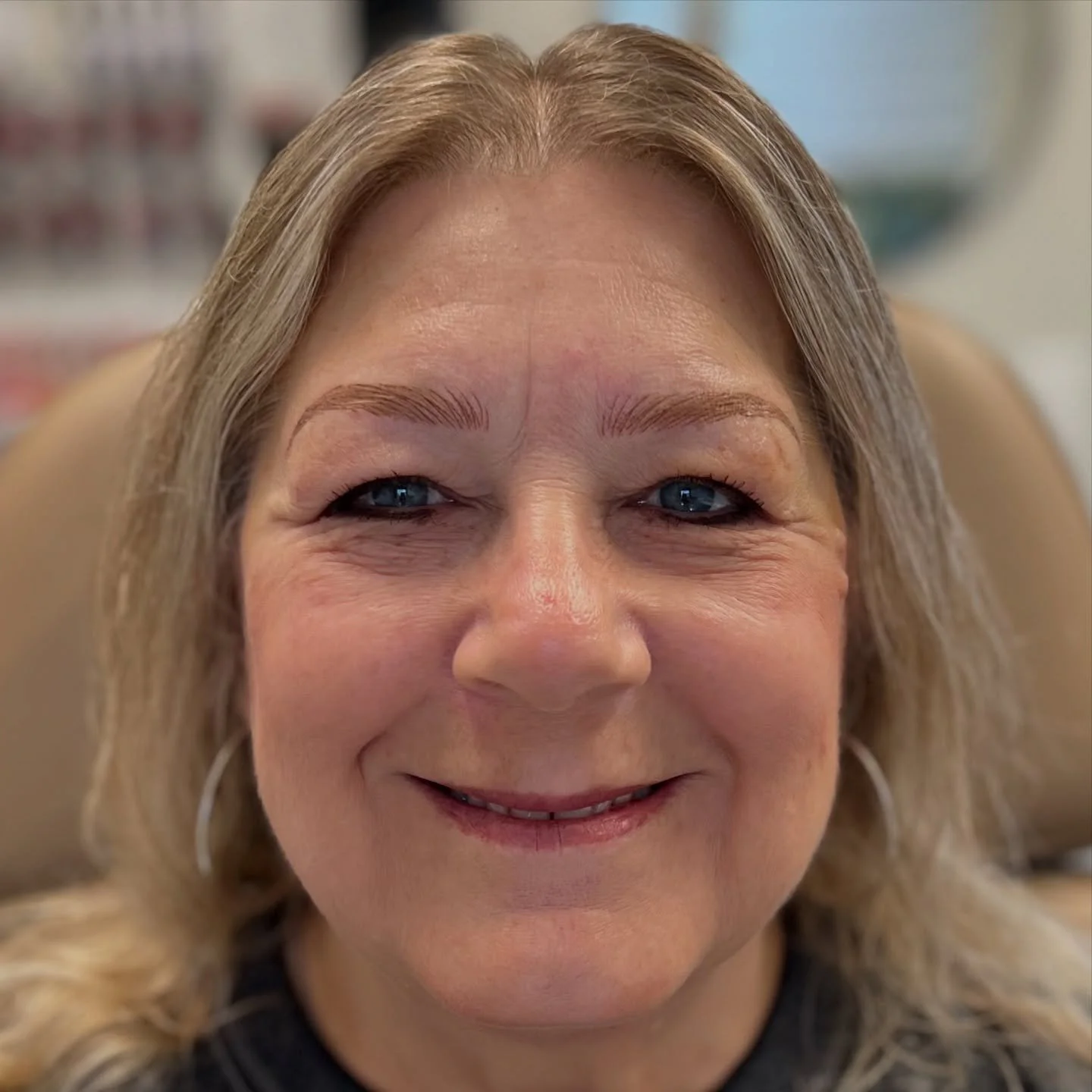 Close-up of a smiling woman with blonde hair, blue eyes, and hoop earrings, sitting in a dental chair.