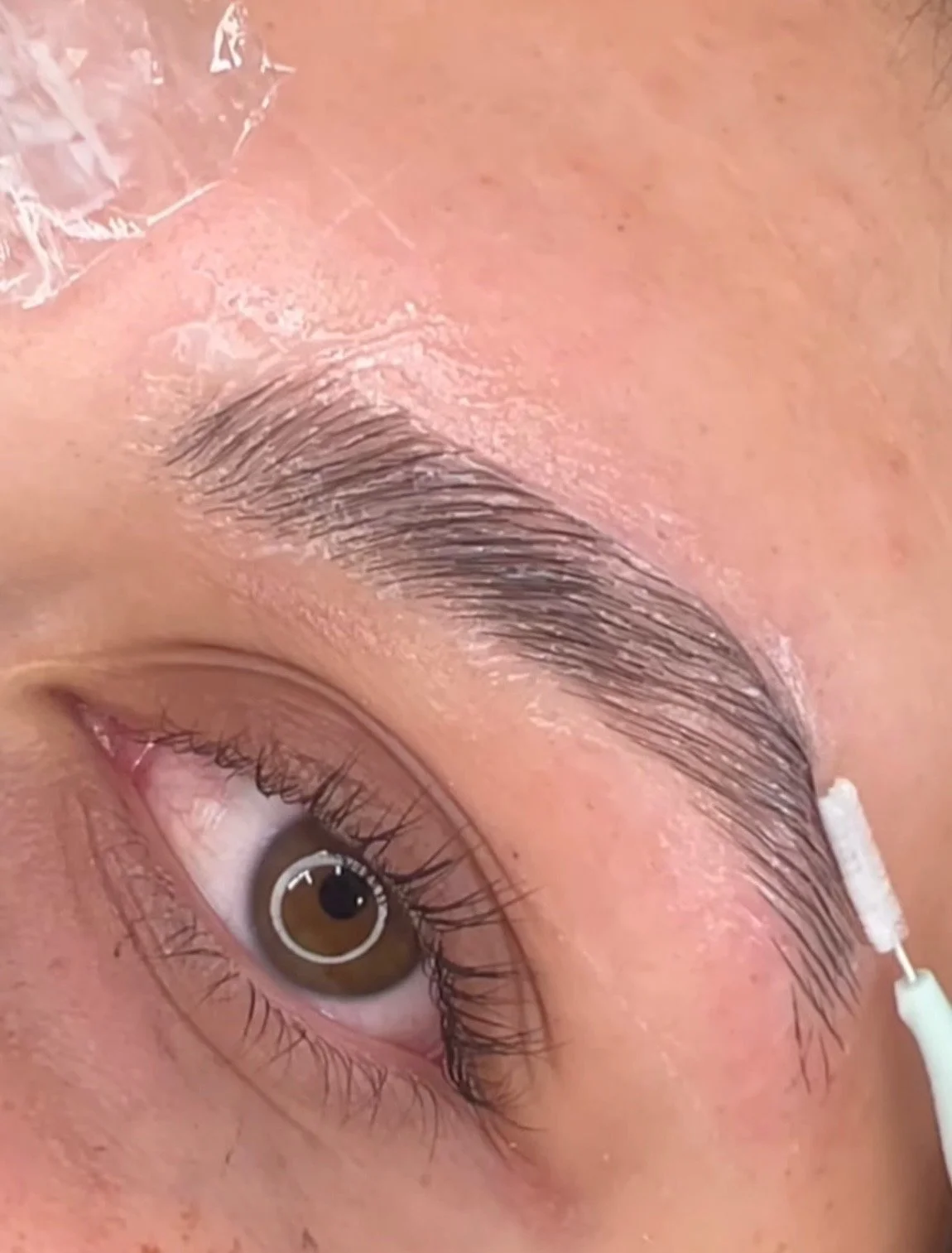 Close-up of a person with brown eyes, applying skincare or makeup with a cotton swab near their eyebrows.