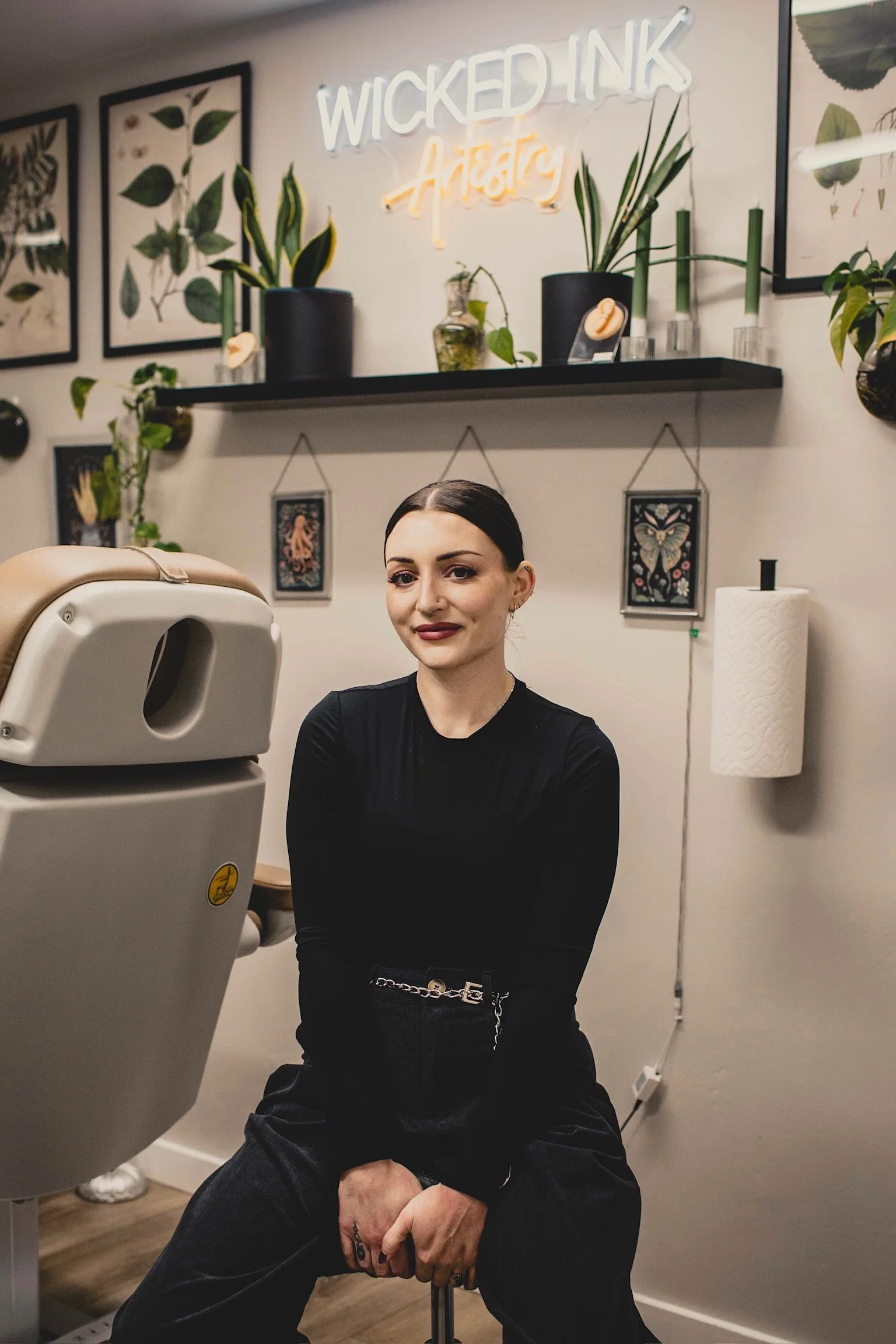 A woman sitting in a tattoo studio with a tattoo machine in front of her. The studio has art prints and potted plants on the wall, including a neon sign that says 'Wicked Ink Artistry'.