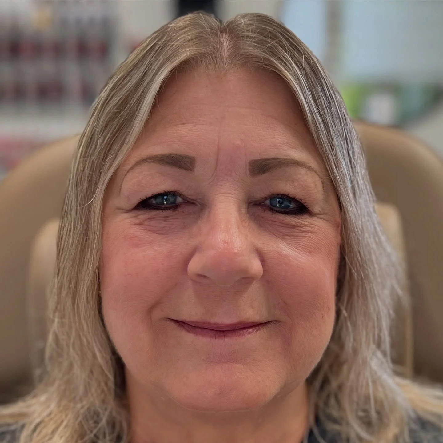 A close-up of a middle-aged woman with blonde hair, blue eyes, and a gentle smile seated in a room with shelves in the background.