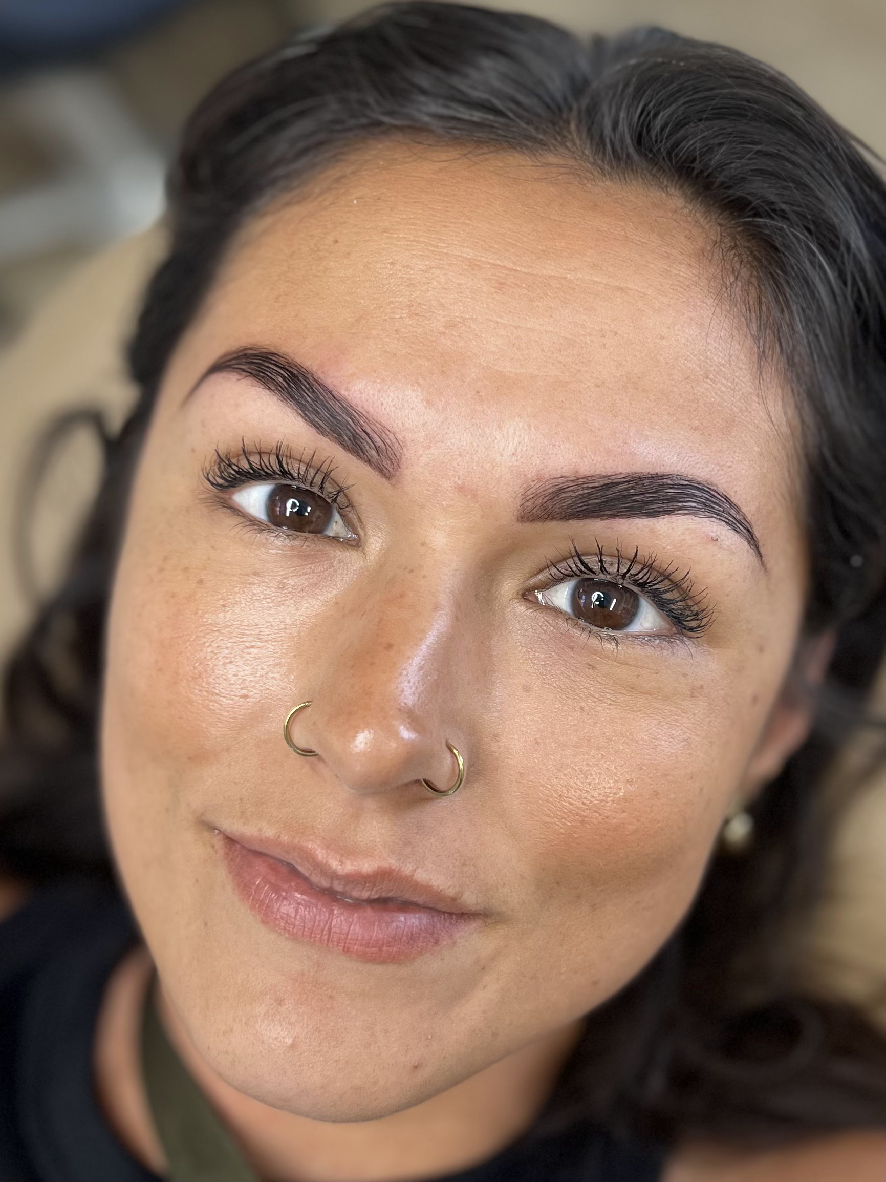 Close-up of a woman with dark hair, brown eyes, well-groomed eyebrows, and a nose piercing, smiling gently.