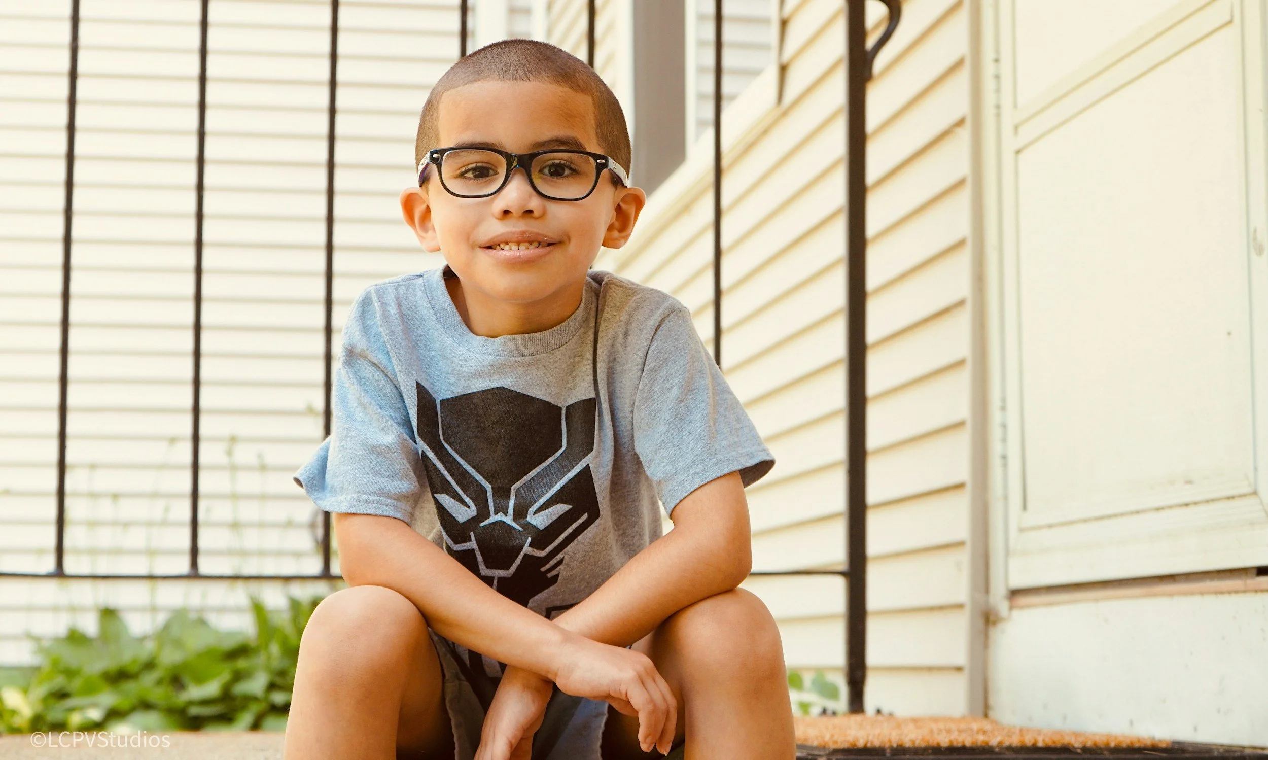 A young boy with glasses and a buzz cut smiling while sitting outside near a house with beige siding and a white door.