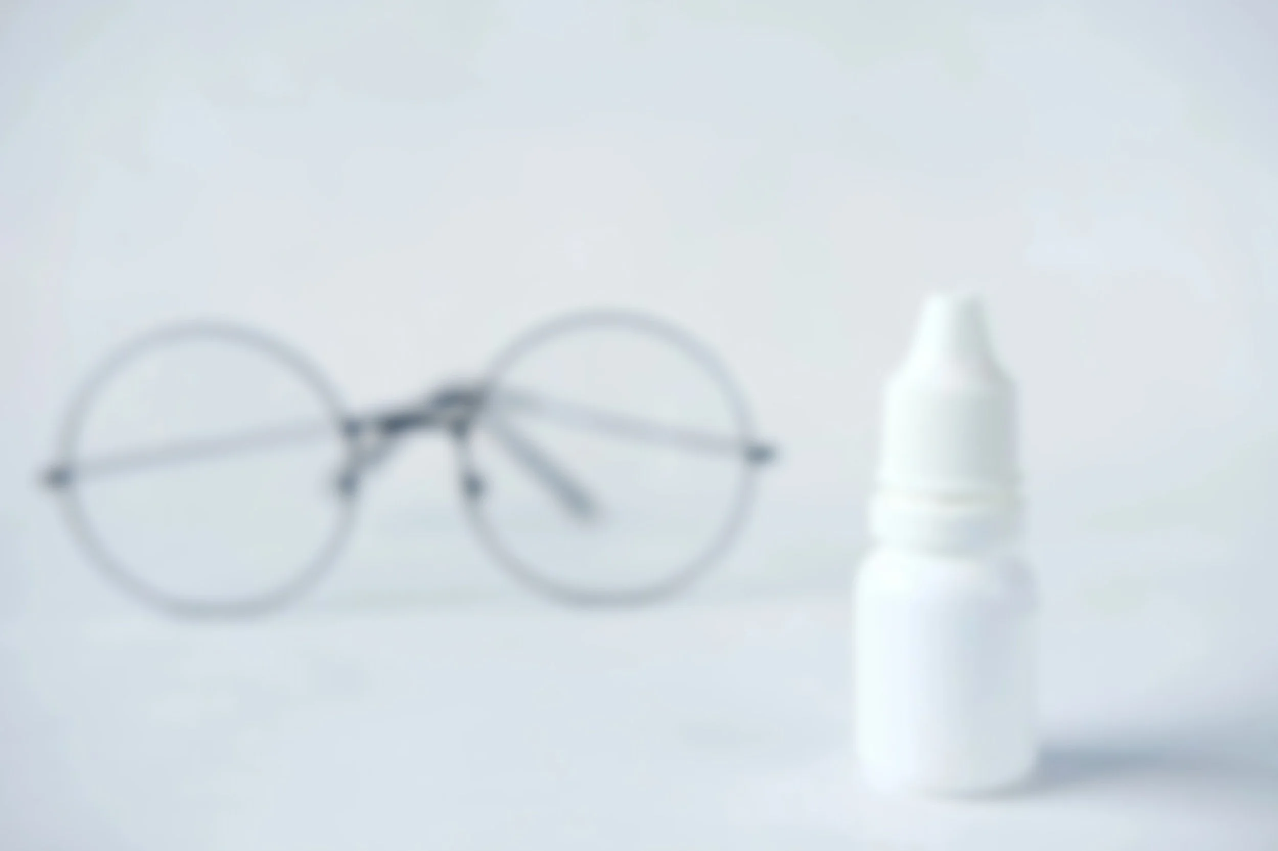 Close-up of eyeglasses, a white bottle, and possibly a small container on a white surface.