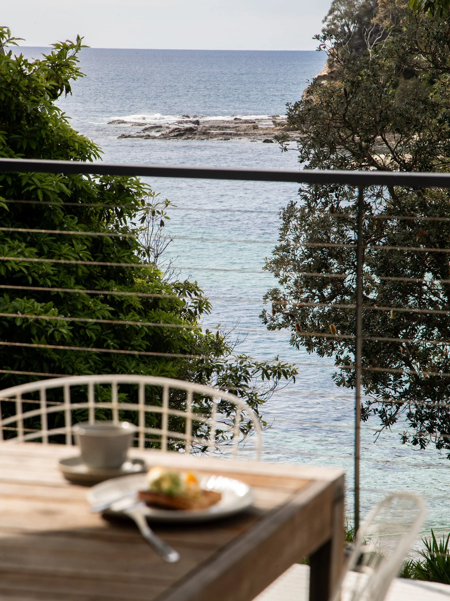 A blurred view of a seaside landscape with clear water, rocks, and foliage, viewed from a porch or deck with a dining table and a cup and plate of food in the foreground.