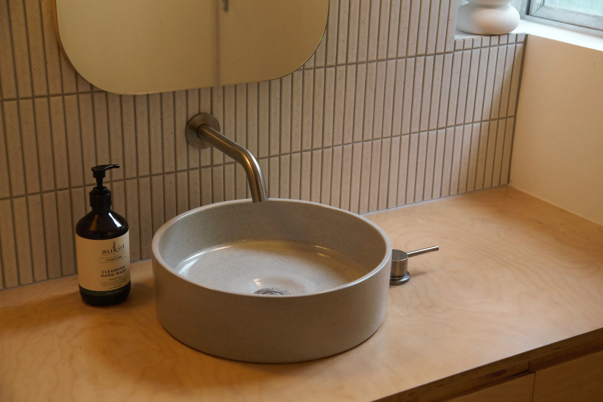 A rounded ceramic sink with a metallic faucet on a light wooden countertop, a bottle of cleansing hand wash labeled 'Sukin' beside it, a circular mirror above, beige tiled wall, and a window with a white vase on the window ledge.