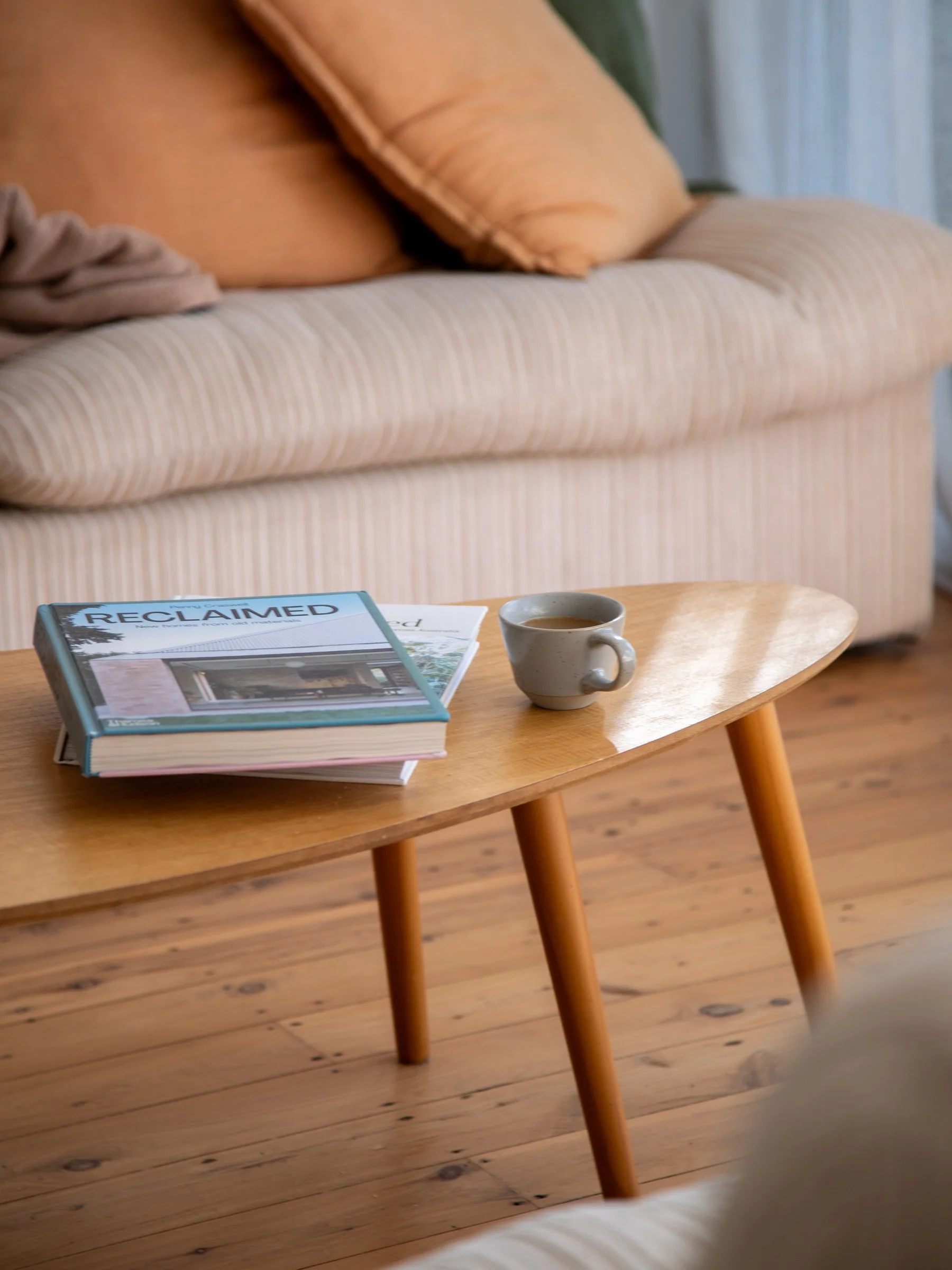 A small wooden table with a stack of books, a coffee mug, and a blanket on a beige armchair in a cozy living room.