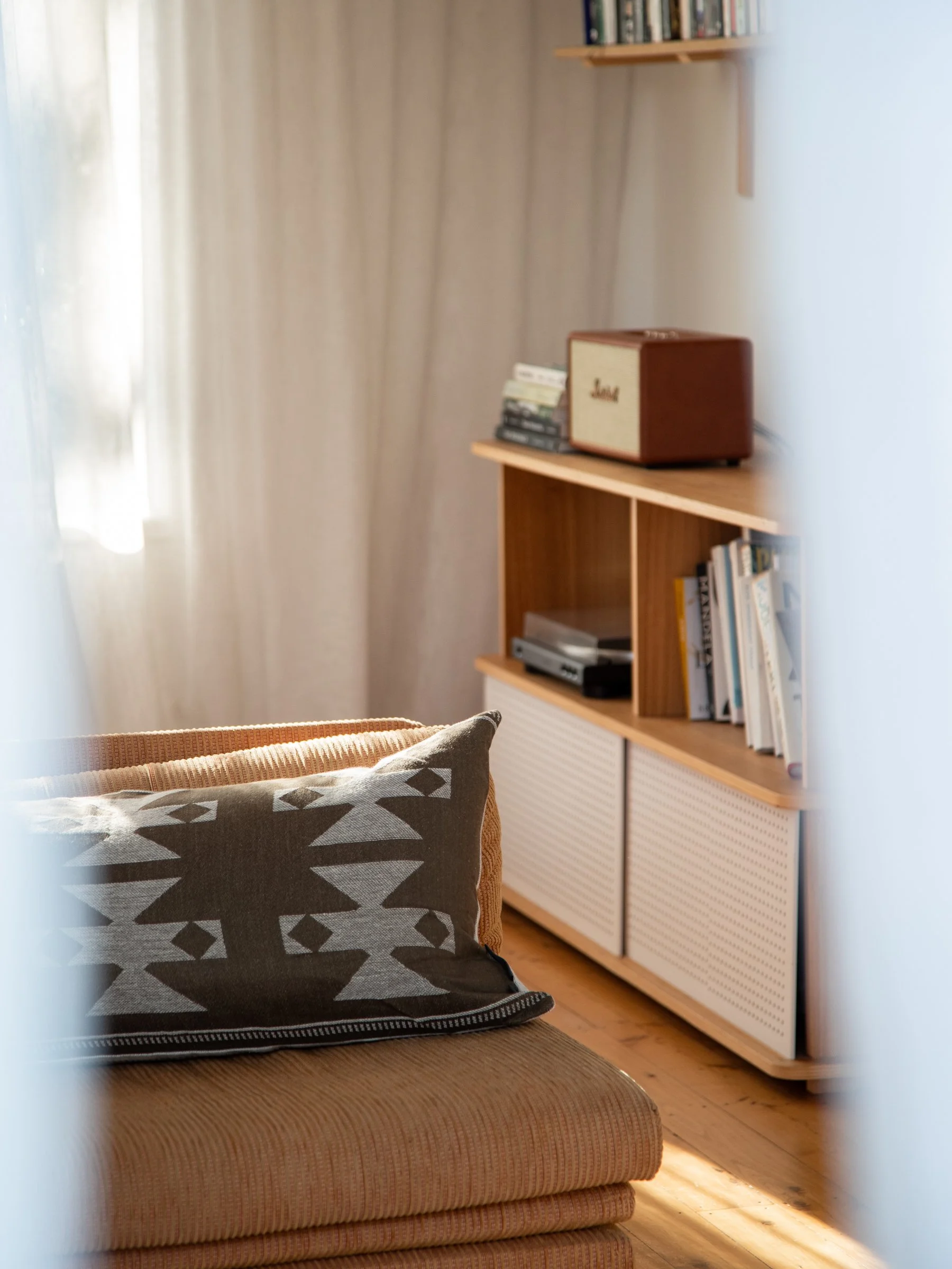 A room with a beige couch, a pillow with a geometric pattern, a wooden shelf with books, and a wooden cabinet with musical equipment.