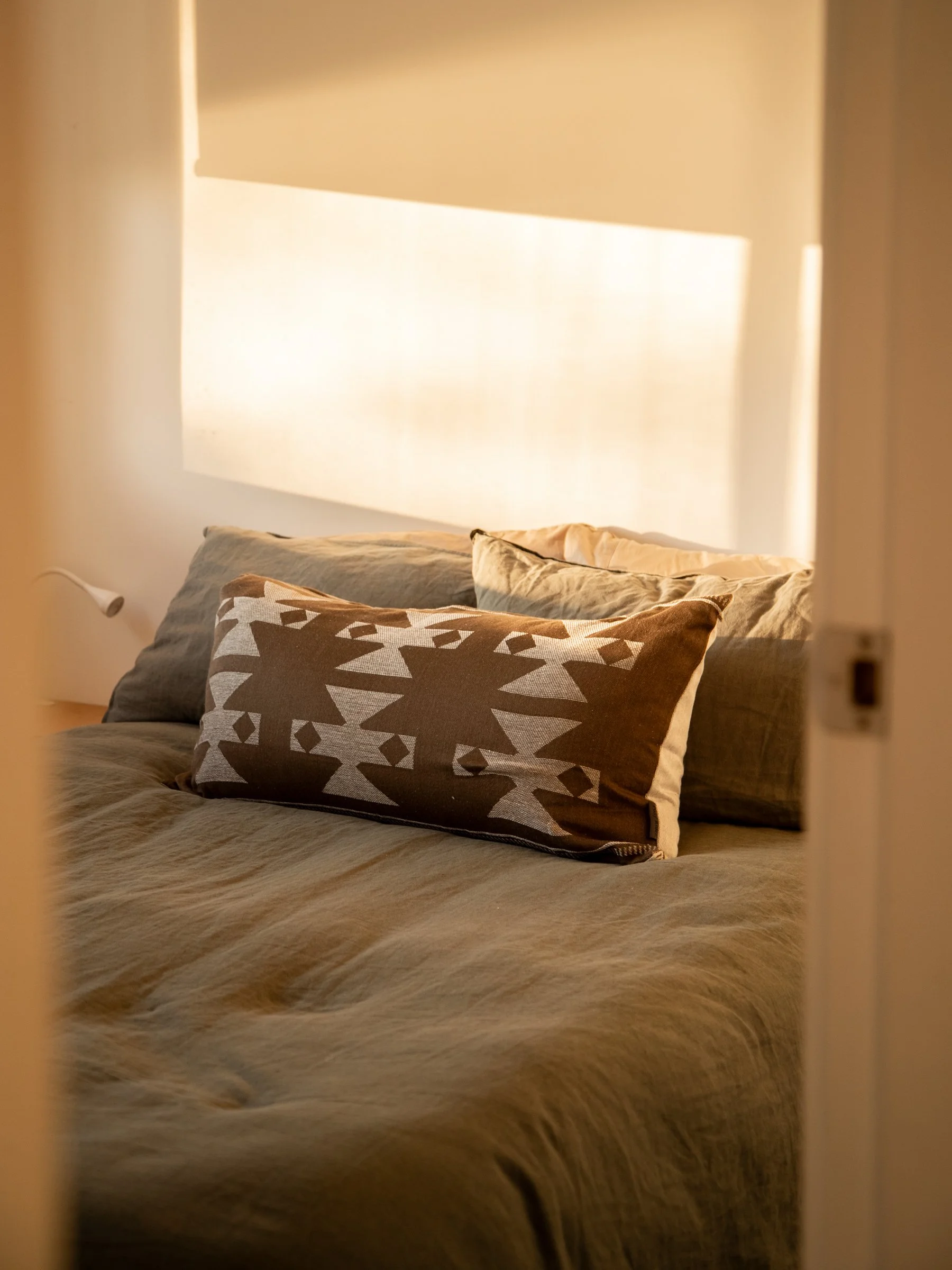 Close-up view of a bed with beige bedding and decorative pillows, viewed through a narrow door or window frame, with sunlight casting shadows on the wall.