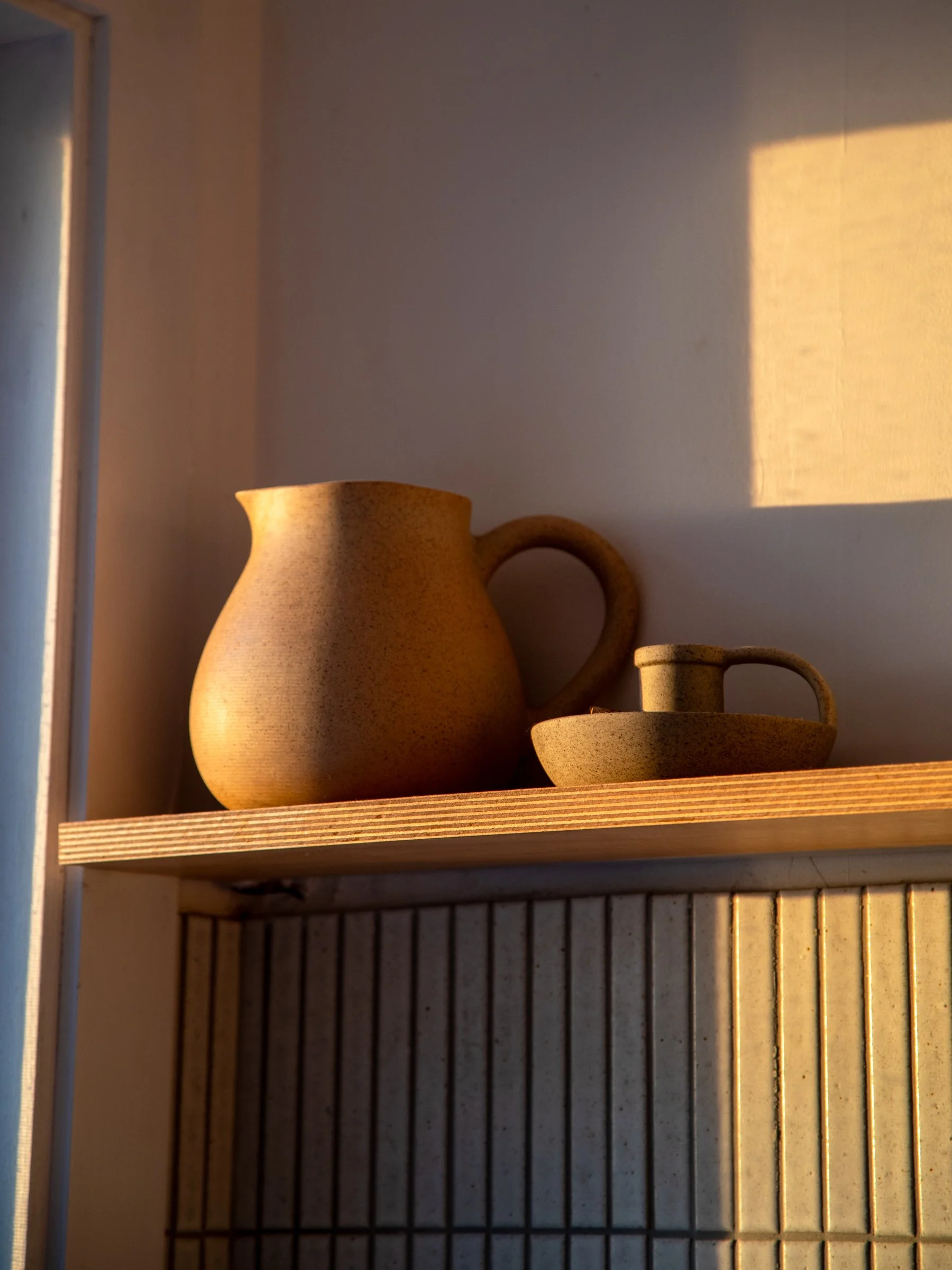 Ceramic pitcher and cup on a wooden shelf inside a cabinet, illuminated by sunlight.