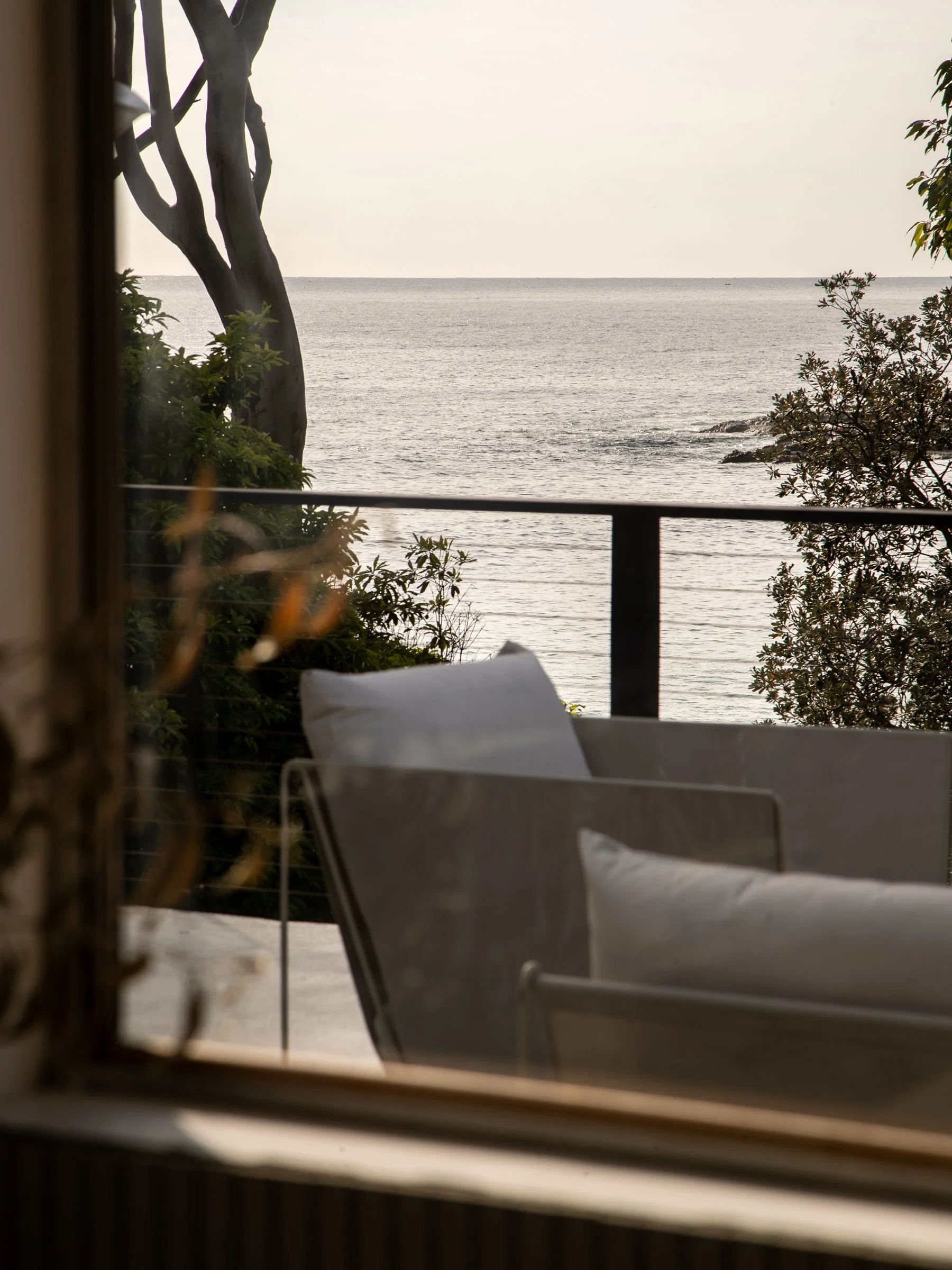 View of the ocean through a window with trees and plants in the foreground, a balcony railing, and a chair with a pillow inside.