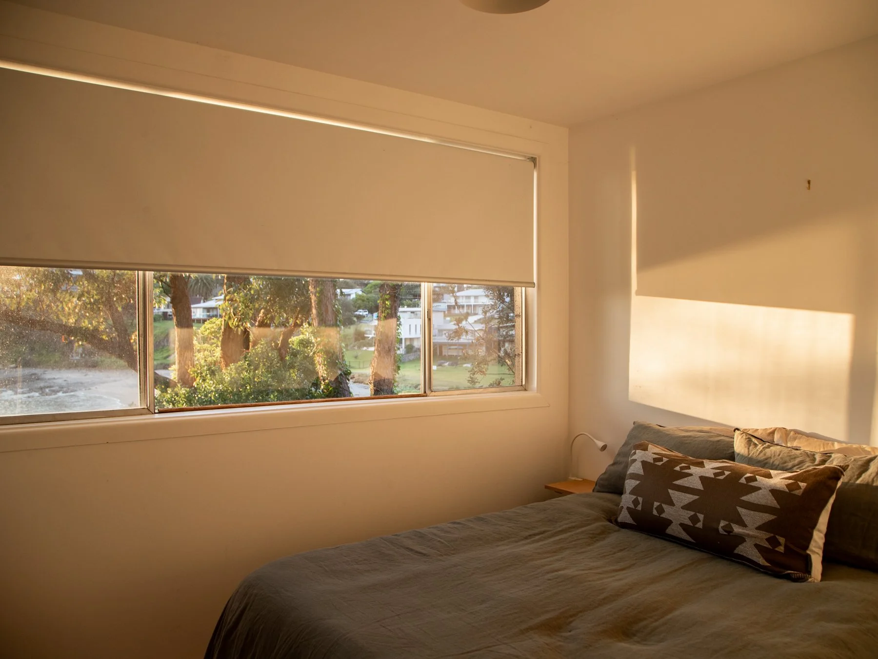 Sunlight coming through a large window with beige blinds, illuminating a bedroom with a bed, pillows, and a small bedside table.