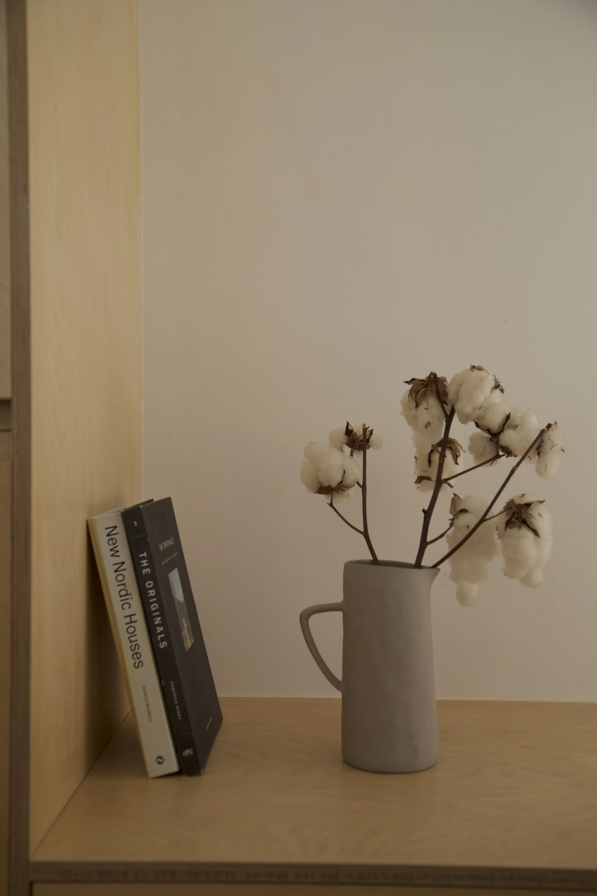 A beige ceramic vase filled with cotton branches sits on a wooden shelf. Two books rest against the side of the shelf titled "New Nordic Houses" and "The Original Houses."