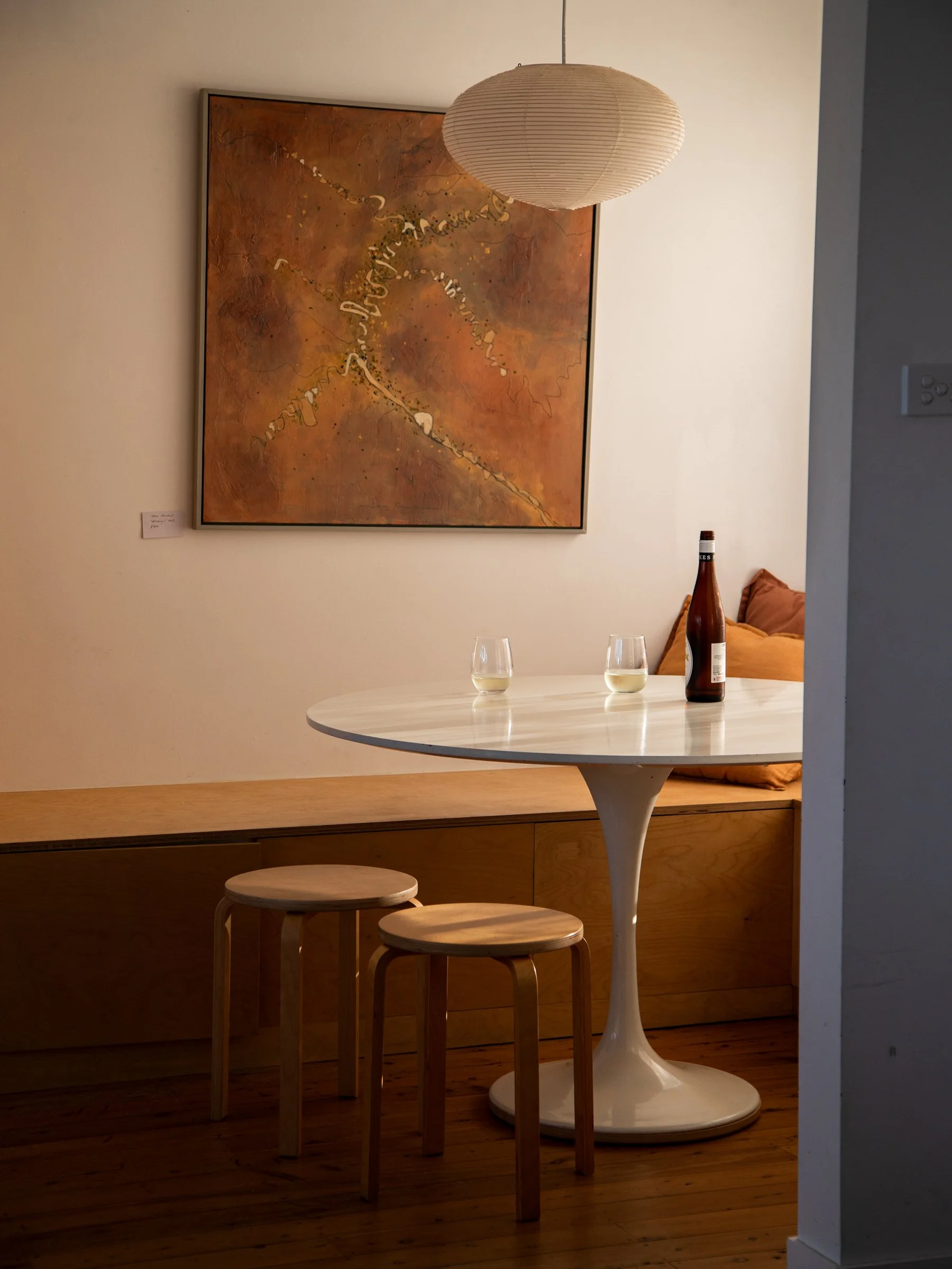A white round table with three wooden stools, a bottle, and two glasses of white wine. There is a wall with a large abstract painting and a paper lantern light fixture.