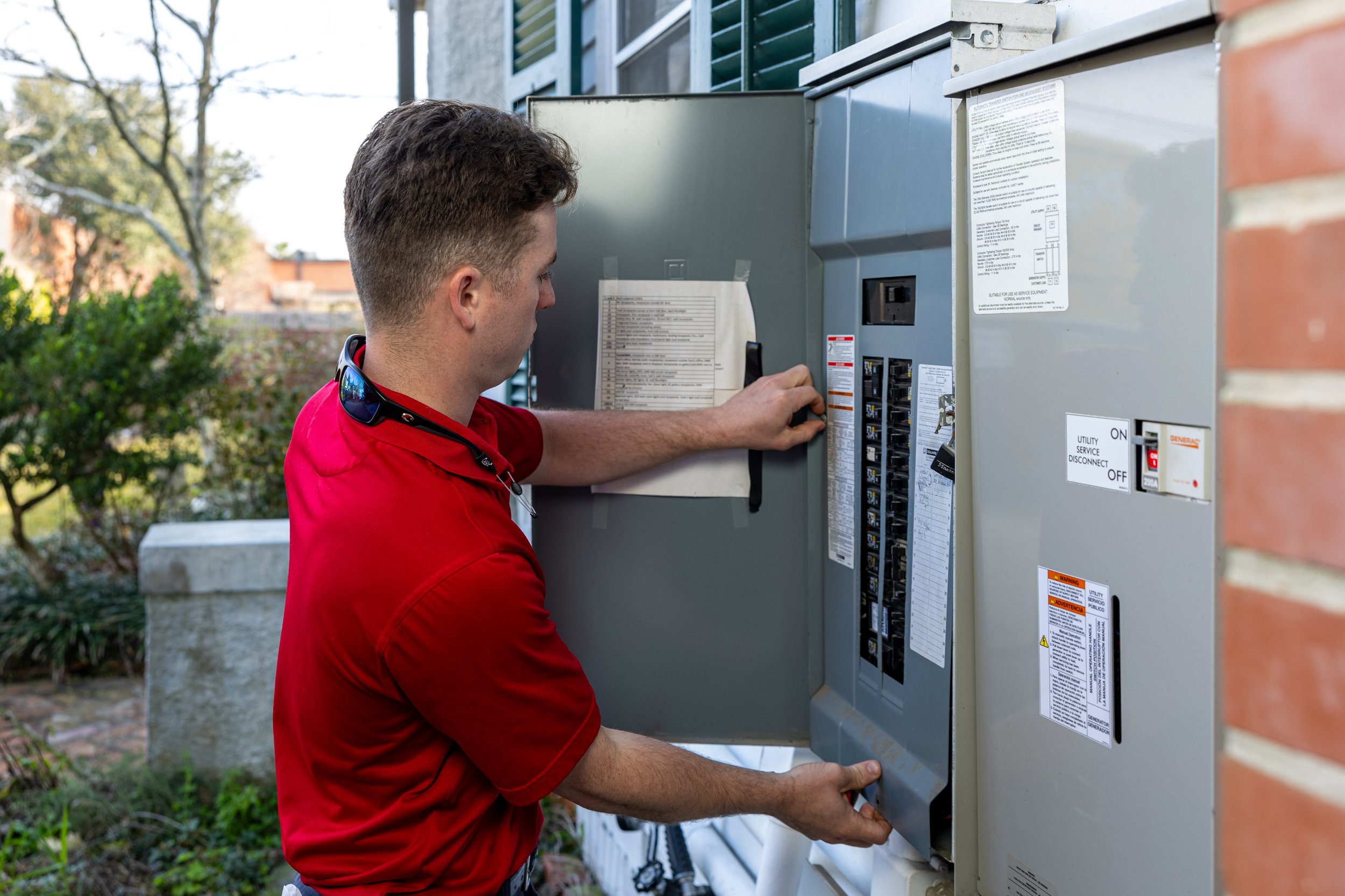A technician in a red shirt working on an outdoor electrical panel, opening the panel door and inspecting the circuit breakers.