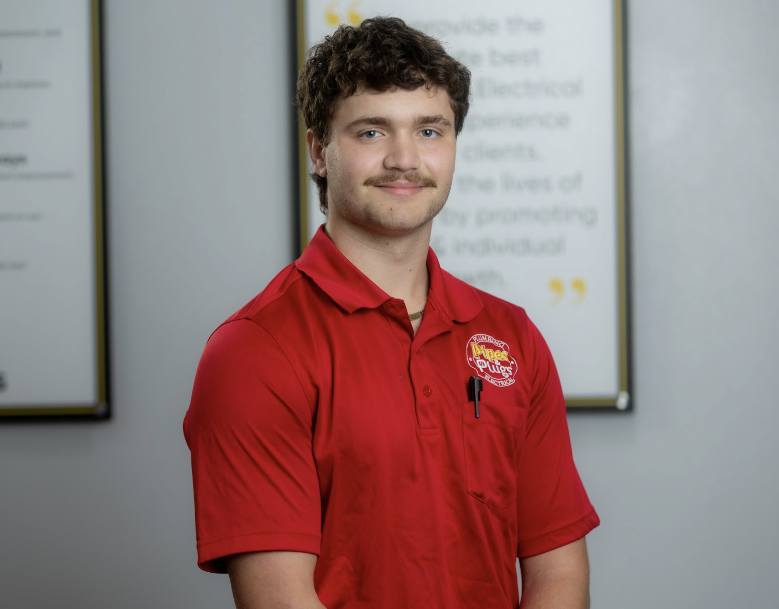 A young man with curly brown hair, mustache, wearing a red uniform shirt with a logo, standing in front of informational posters.