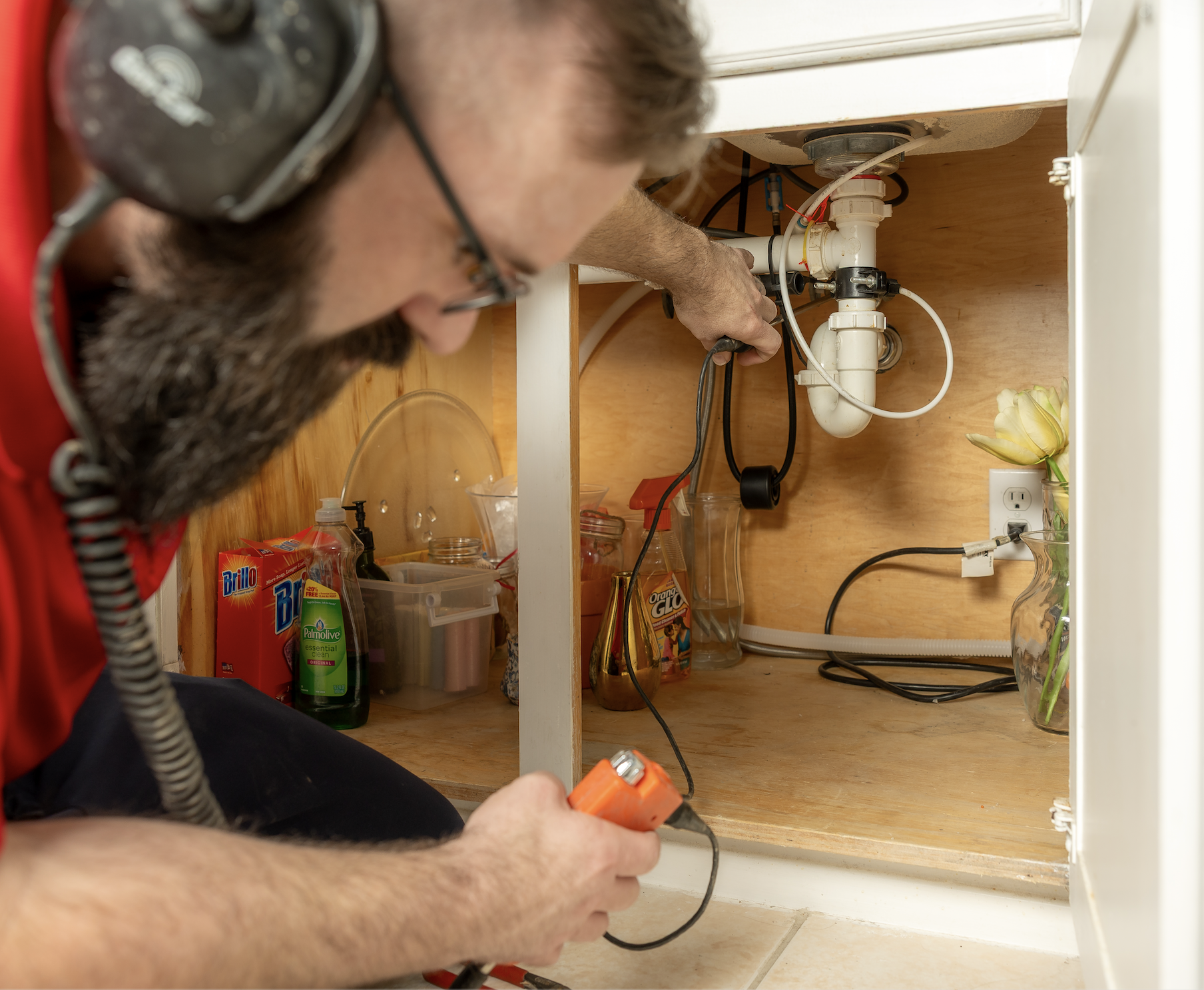 A man working on the plumbing underneath a kitchen sink, using a tool while wearing protective ear muffs. The cabinet has various cleaning supplies and a vase with flowers.