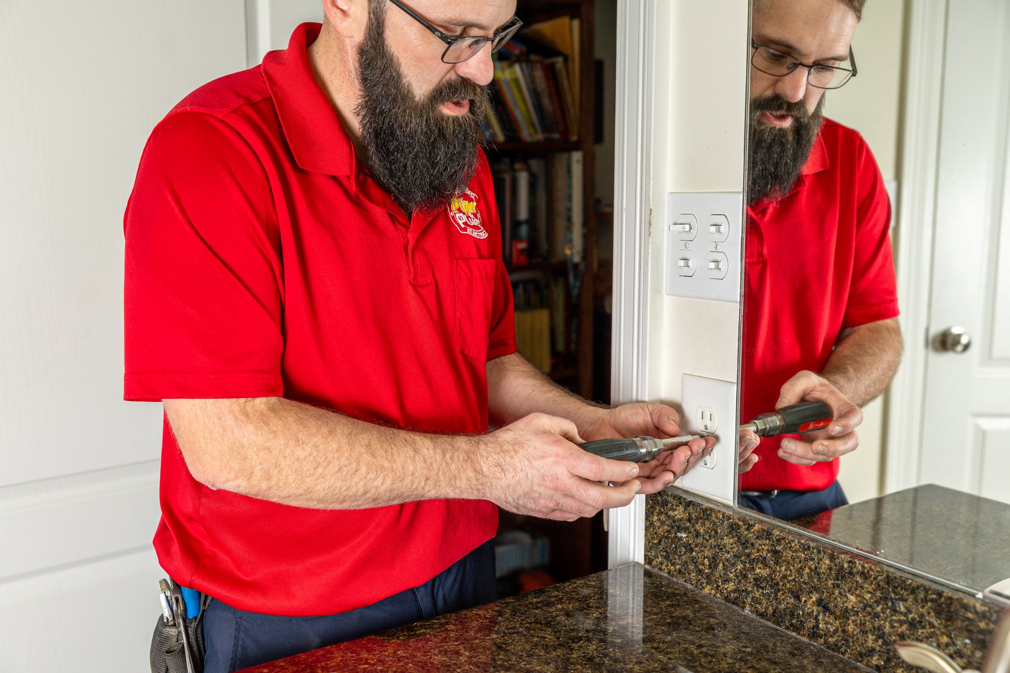 A man with a beard and glasses, wearing a red shirt, is using a screwdriver to work on an outlet cover in a home kitchen or bathroom. His reflection is visible in a mirror on the wall.