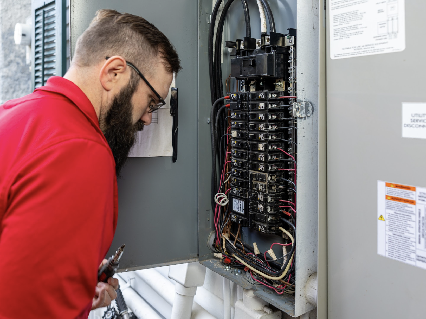 A technician inspecting electrical wiring inside an open utility electrical panel, wearing a red uniform.