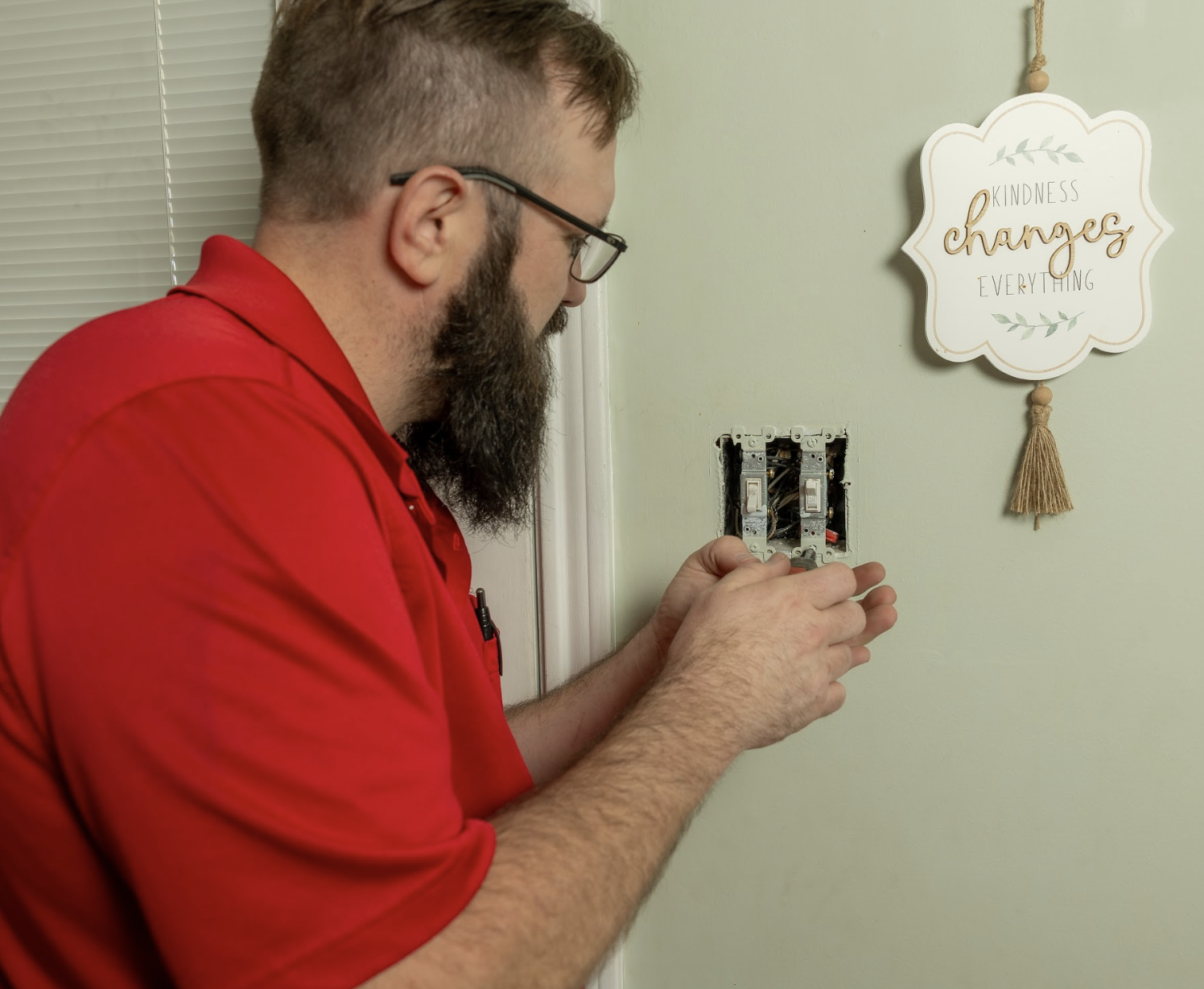 A man in a red shirt with glasses and a beard installing or repairing an electrical switch in a wall beside a decorative wall hanging that says 'Kindness changes everything'.