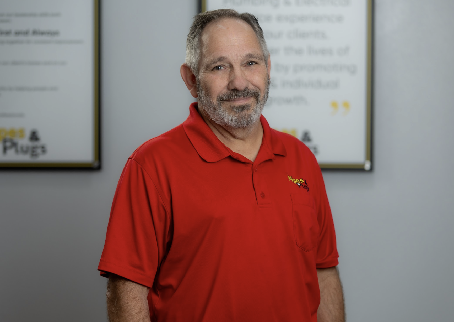 A middle-aged man with gray hair and beard, smiling, wearing a red polo shirt with a logo, standing indoors in front of blurred posters.