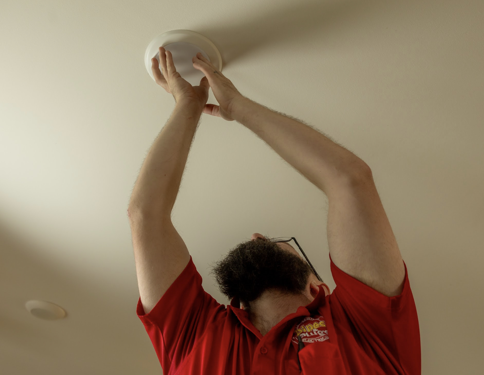 A man wearing a red shirt and glasses is changing a ceiling light fixture.