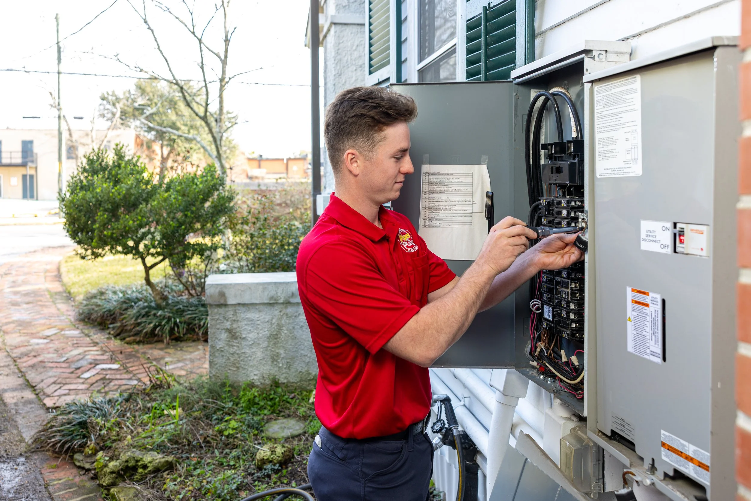 A male technician in a red uniform inspecting an electrical panel outside a building.