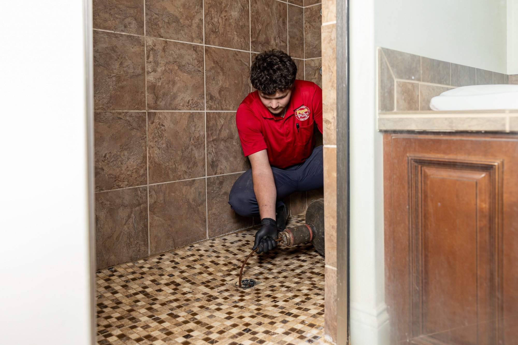A plumber in a red uniform working on a bathroom drain in a tile shower, kneeling on the floor, using a pipe wrench.