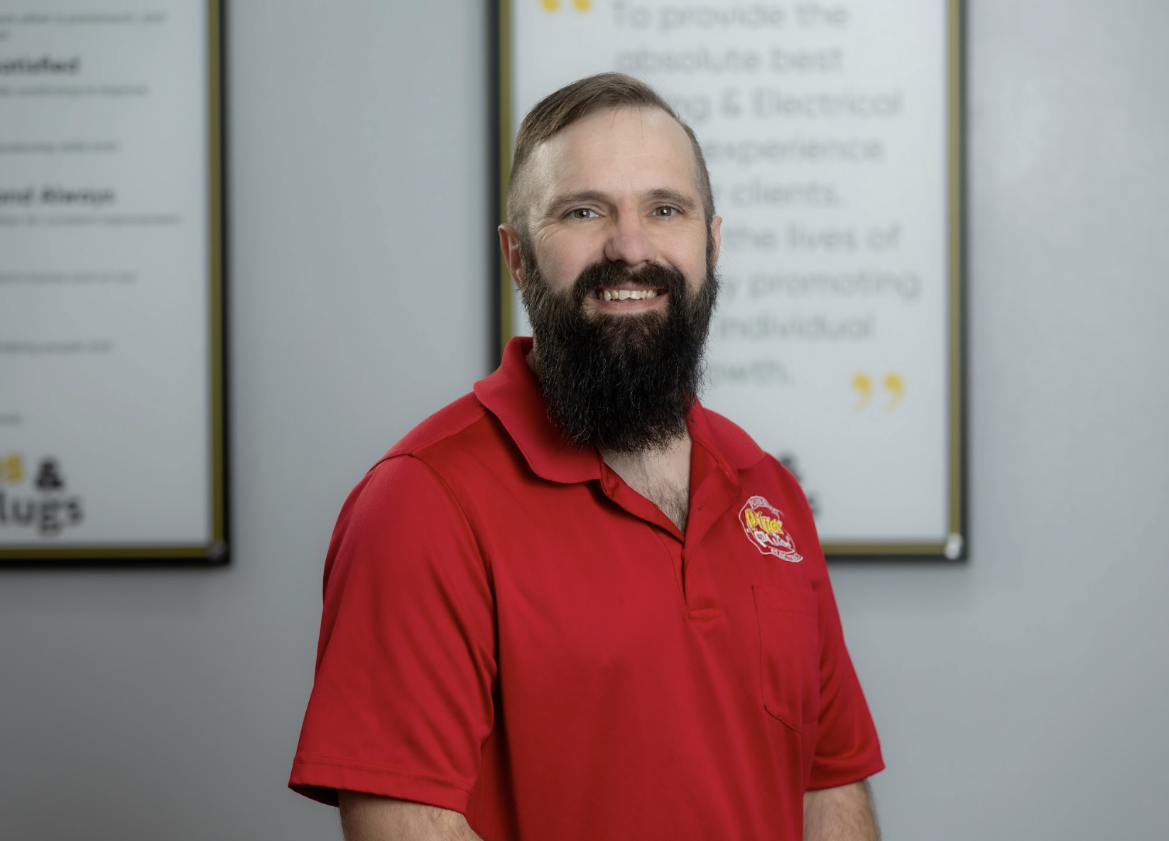 A man with a beard and mustache wearing a red polo shirt, standing in front of a blurred background with framed posters or signs.