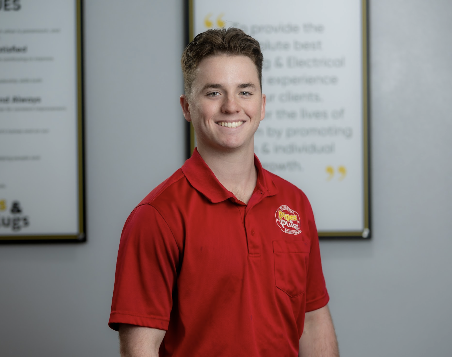 A young man with short brown hair, smiling, wearing a red polo shirt with a company logo, standing indoors in front of framed posters.
