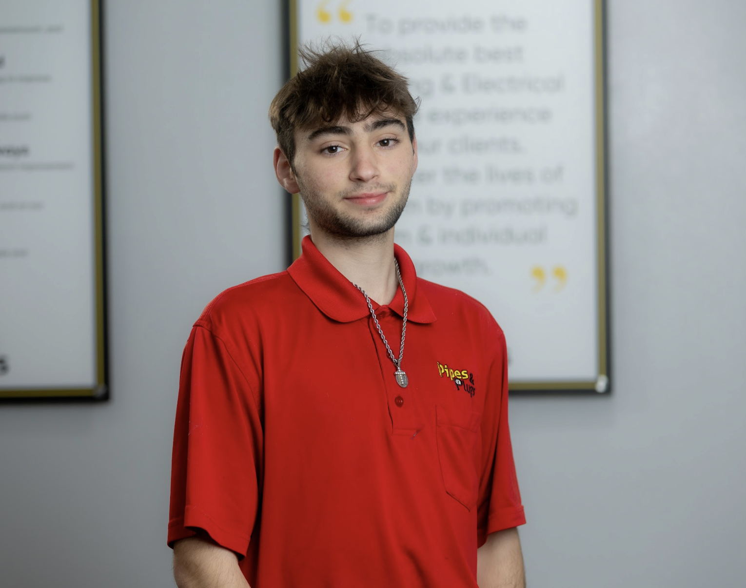A young man with light brown hair and facial hair, wearing a red uniform shirt with a logo on it, standing indoors in front of blurred bulletin boards or signs.