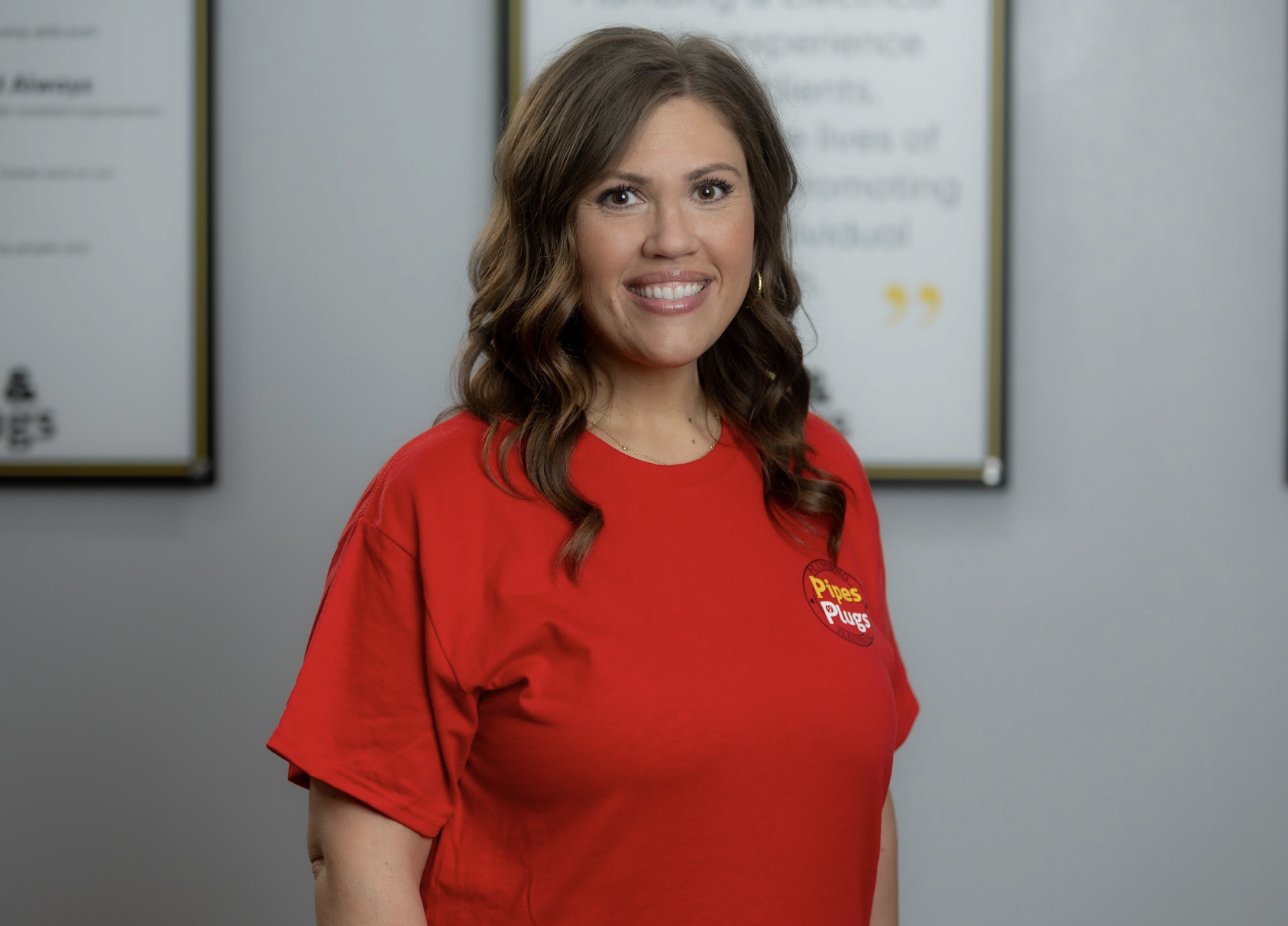 A woman with brown hair wearing a red t-shirt with a 'Pipes & Plugs' logo, smiling in front of white boards with text.