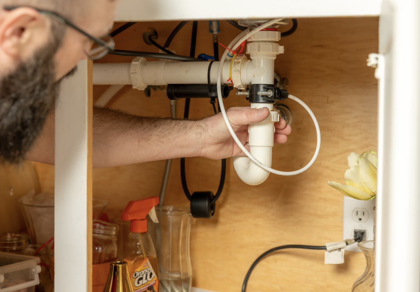 A person repairing or inspecting plumbing pipes under a kitchen sink, with visible PVC pipes, hoses, and an electrical outlet with a plugged-in cord.