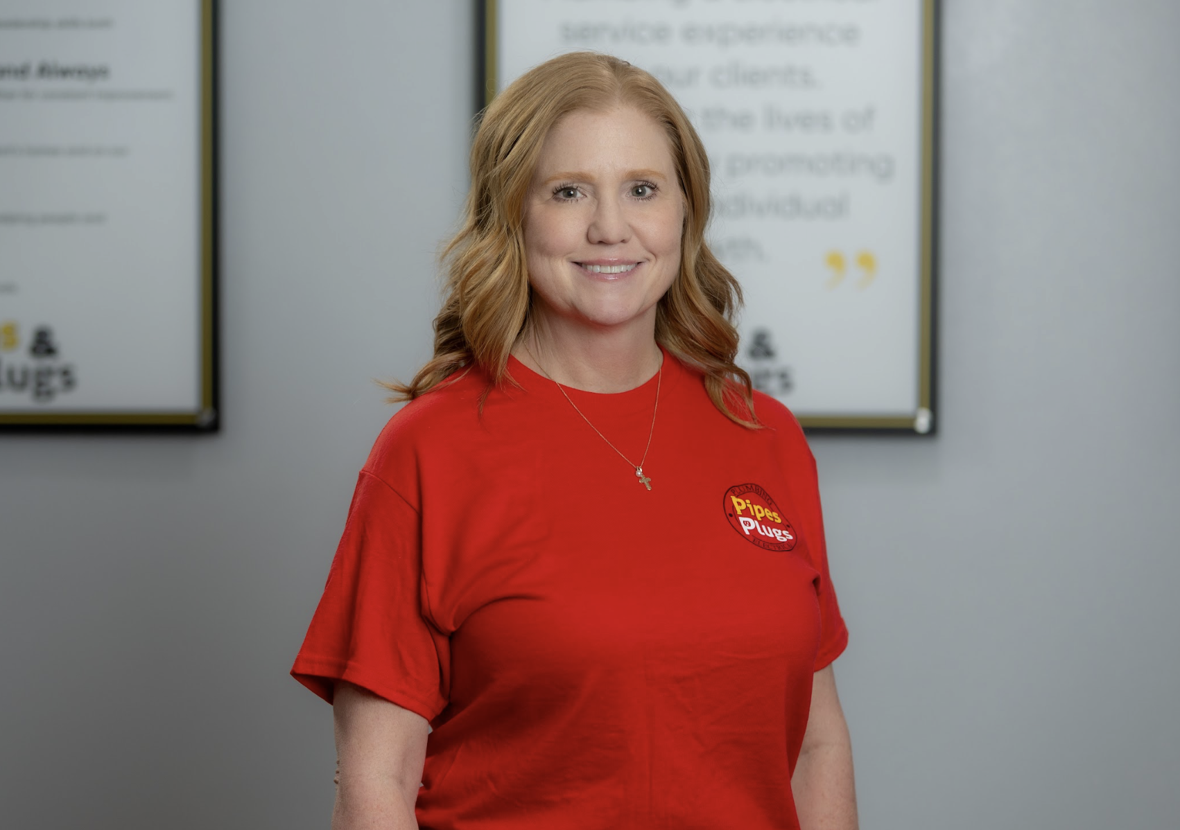 A woman with red hair smiling, wearing a red shirt with a logo that says 'Pipes & Plugs,' standing indoors with framed posters in the background.