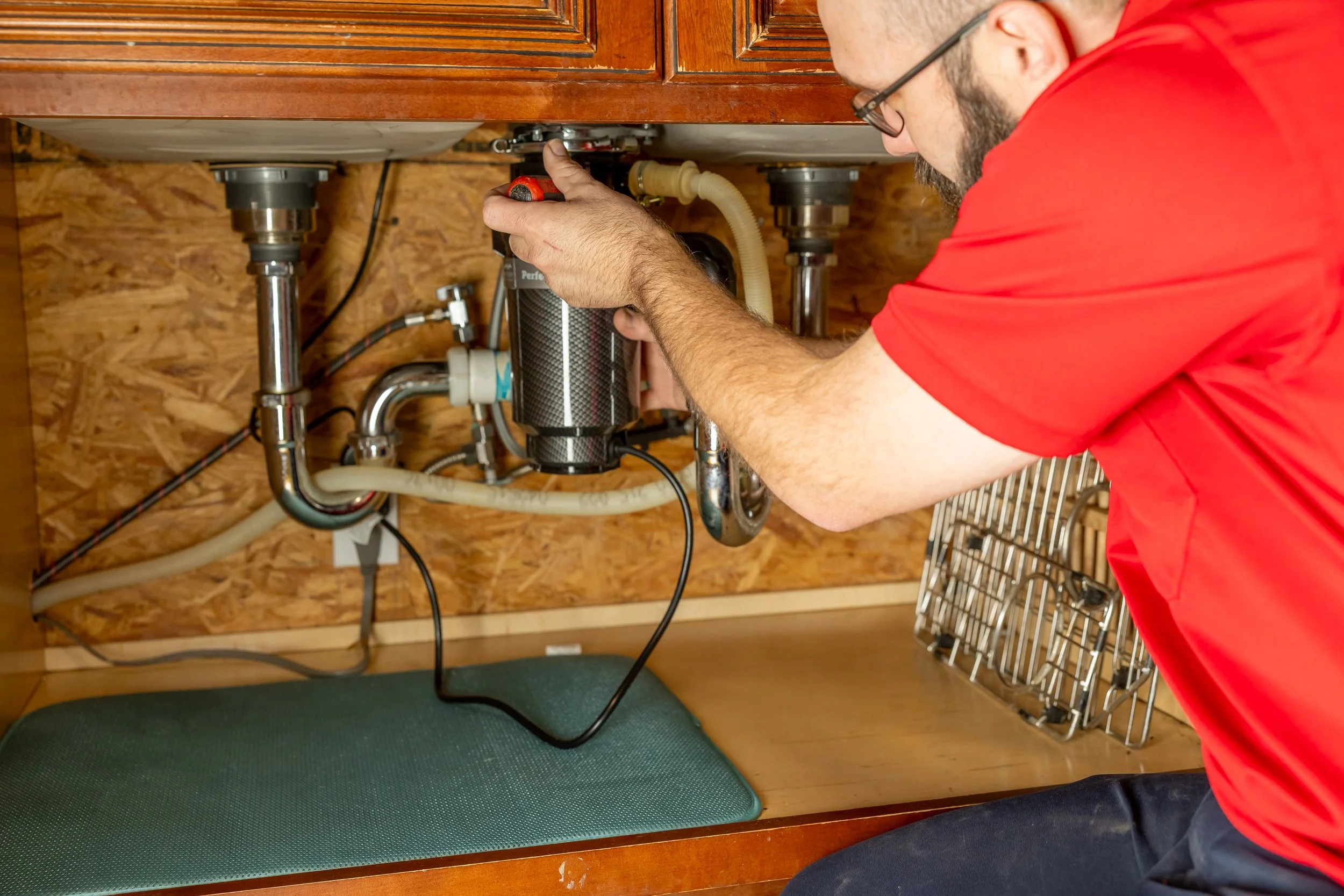 A person in a red shirt working underneath a kitchen sink, inspecting or repairing the plumbing with a tool.
