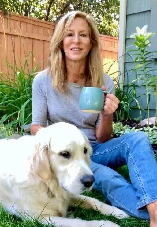 A woman sitting on the grass outdoors next to a large white dog, holding a blue mug and smiling, with greenery and a wooden fence in the background.