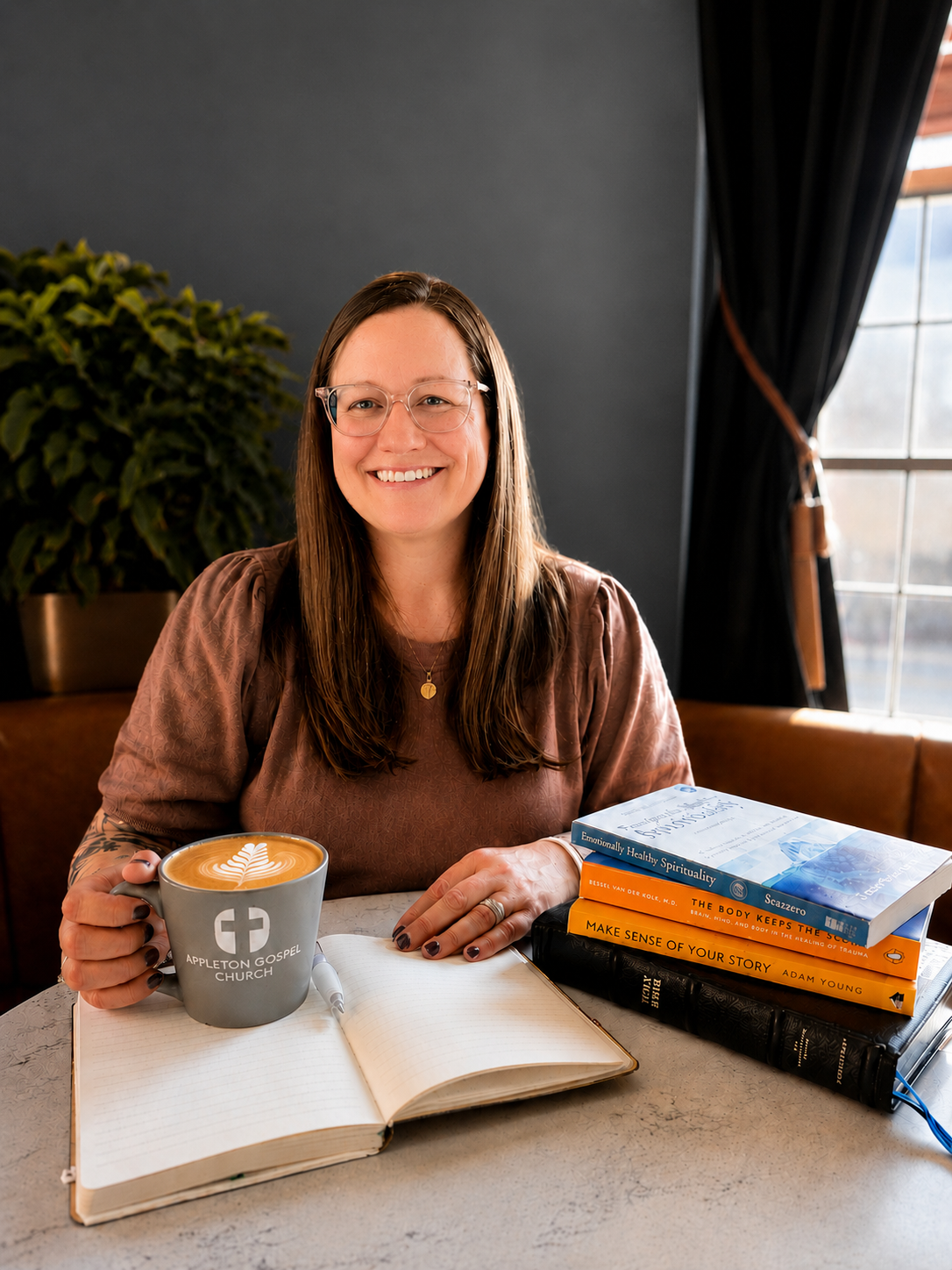 A woman with long brown hair and glasses smiling at the camera, sitting at a table with a latte in a gray mug that says 'Appleton Gospel Church,' an open notebook, and a stack of books, in a room with a large window, dark curtains, and a potted plant in the background.