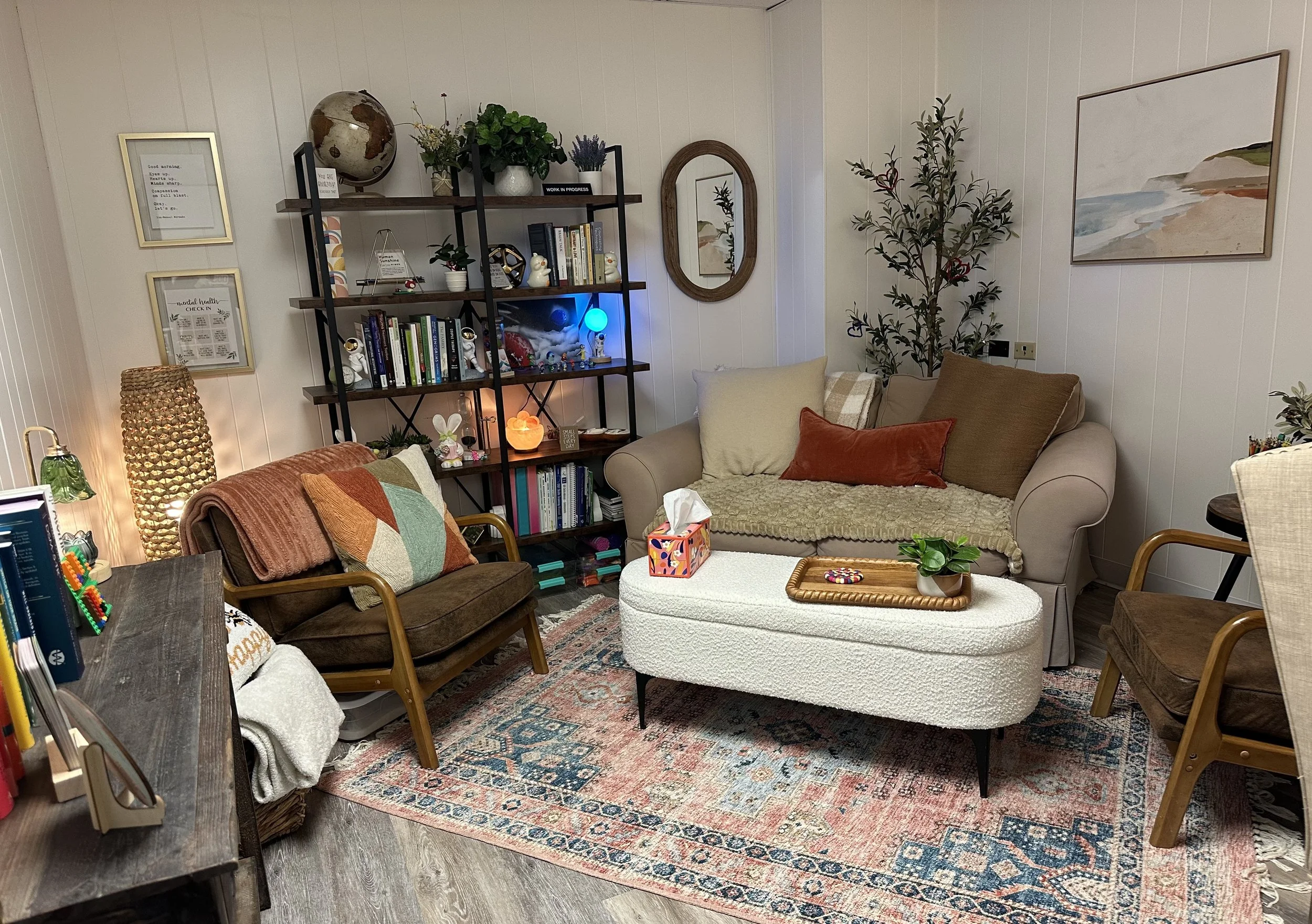 Living room with a white wall, a black metal bookshelf with plants and books, a beige sofa with throw pillows, a colorful rug, a wooden coffee table, and framed artwork and mirror on the wall.
