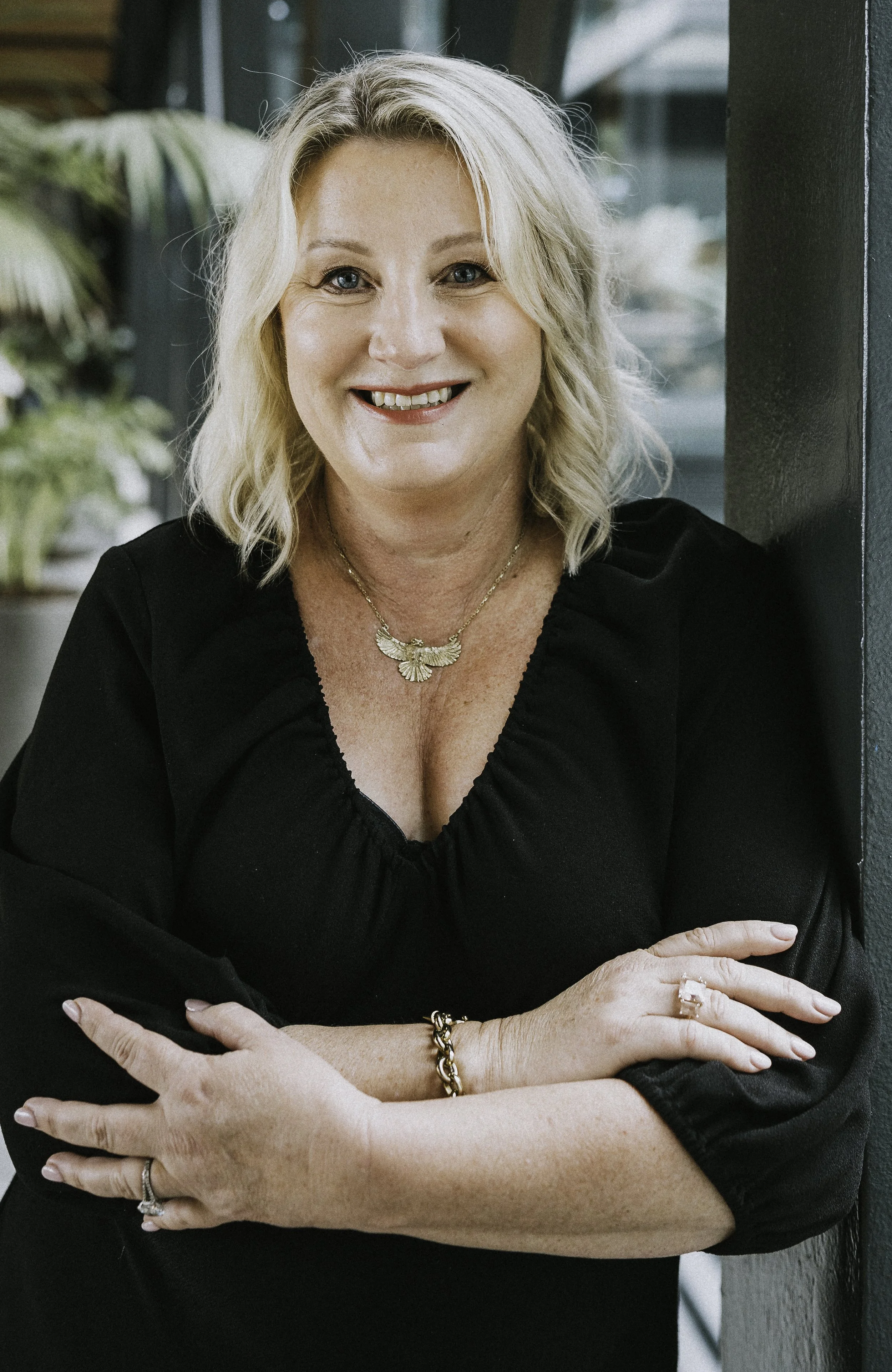 Image of Juanita Neville Te Rito - blonde hair and blue eyes, smiling, wearing a black top, gold jewelry, standing indoors near large windows with plants in the background.
