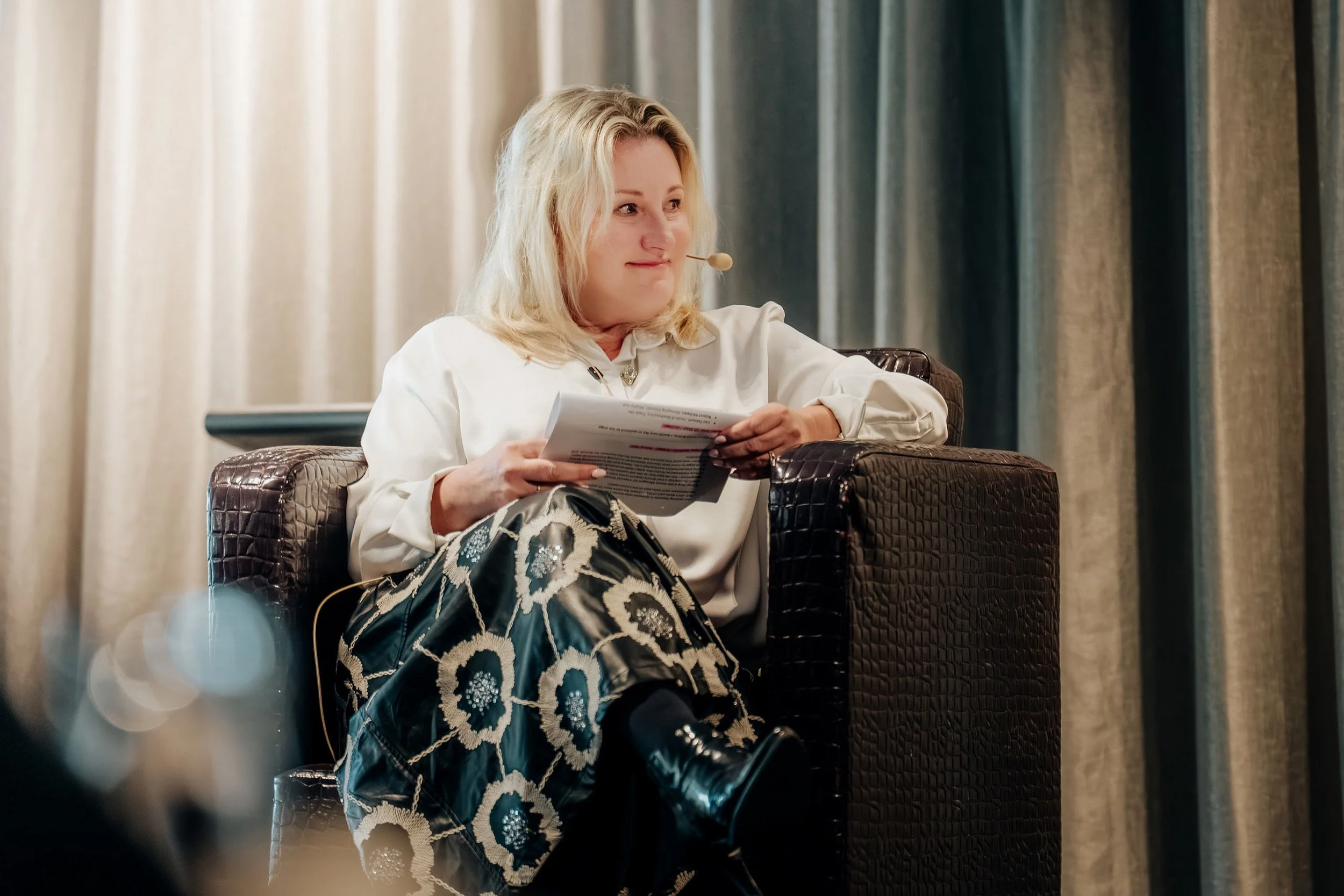 Juanita Neville Te Rito with blonde hair sitting in a black leather armchair, holding a paper, wearing a white blouse and a patterned skirt with black boots, indoors with beige curtains in the background.