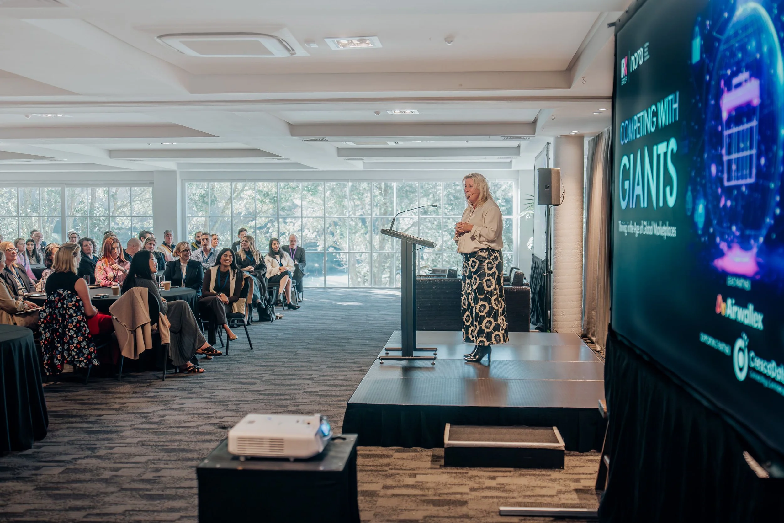Juanita Neville Te Rito giving a presentation on stage in front of a seated audience in a conference room with large windows, a banner with the title 'Competing with Giants,' and a large digital screen.